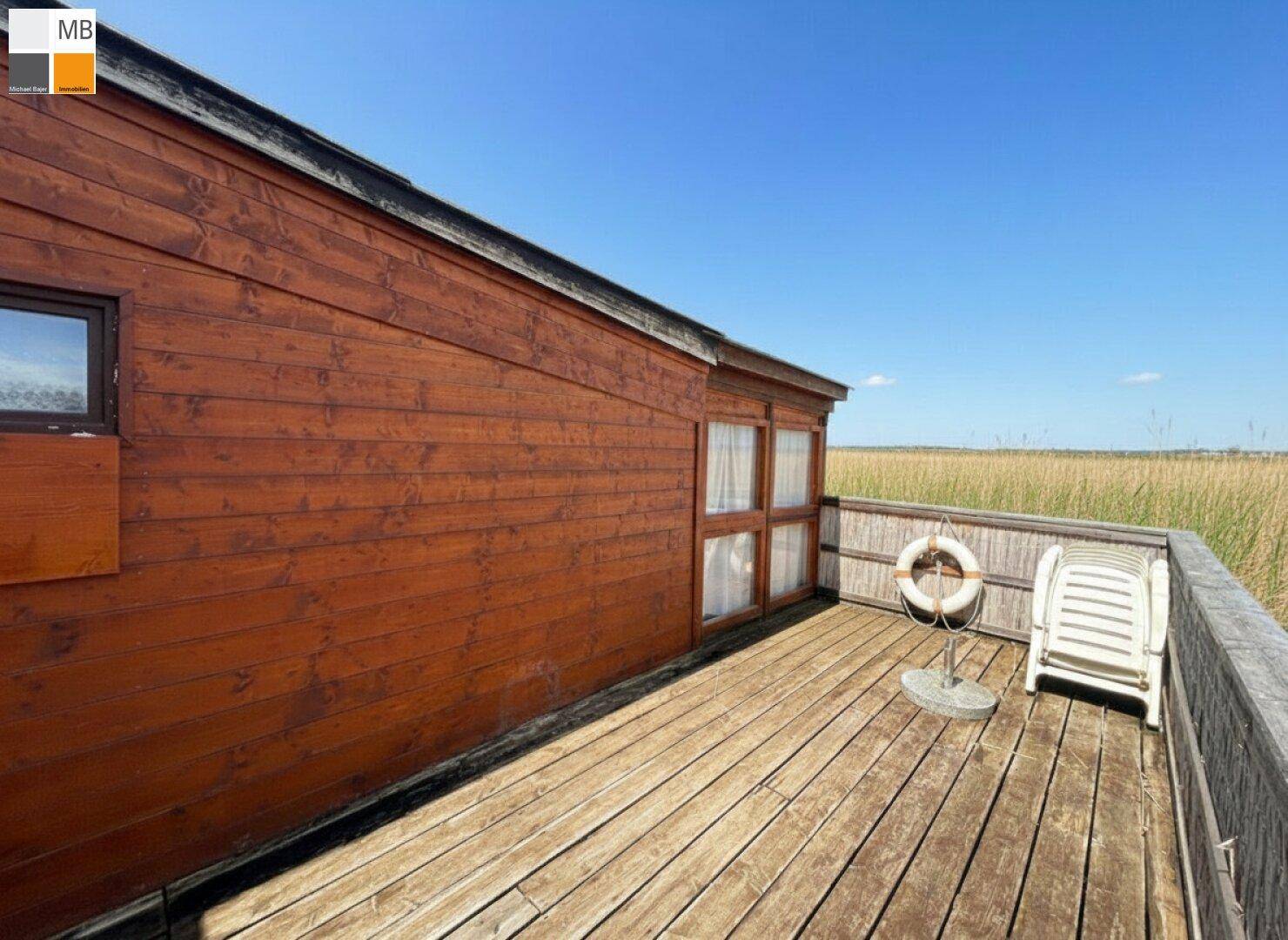 Holzterrasse mit Liegestuhl und Rettungsring, bietet weiten Blick über die Schilflandschaft.