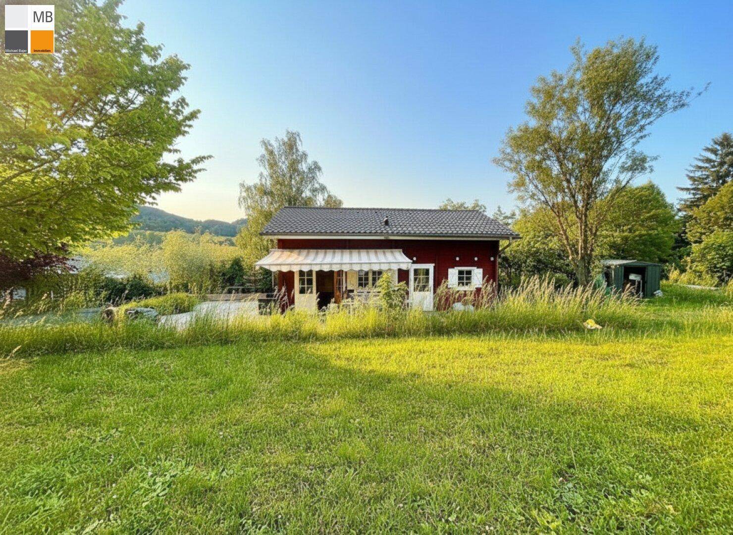 Charmantes rotes Holzhaus mit weißer Markise, umgeben von einer weitläufigen grünen Wiese und Bäumen.