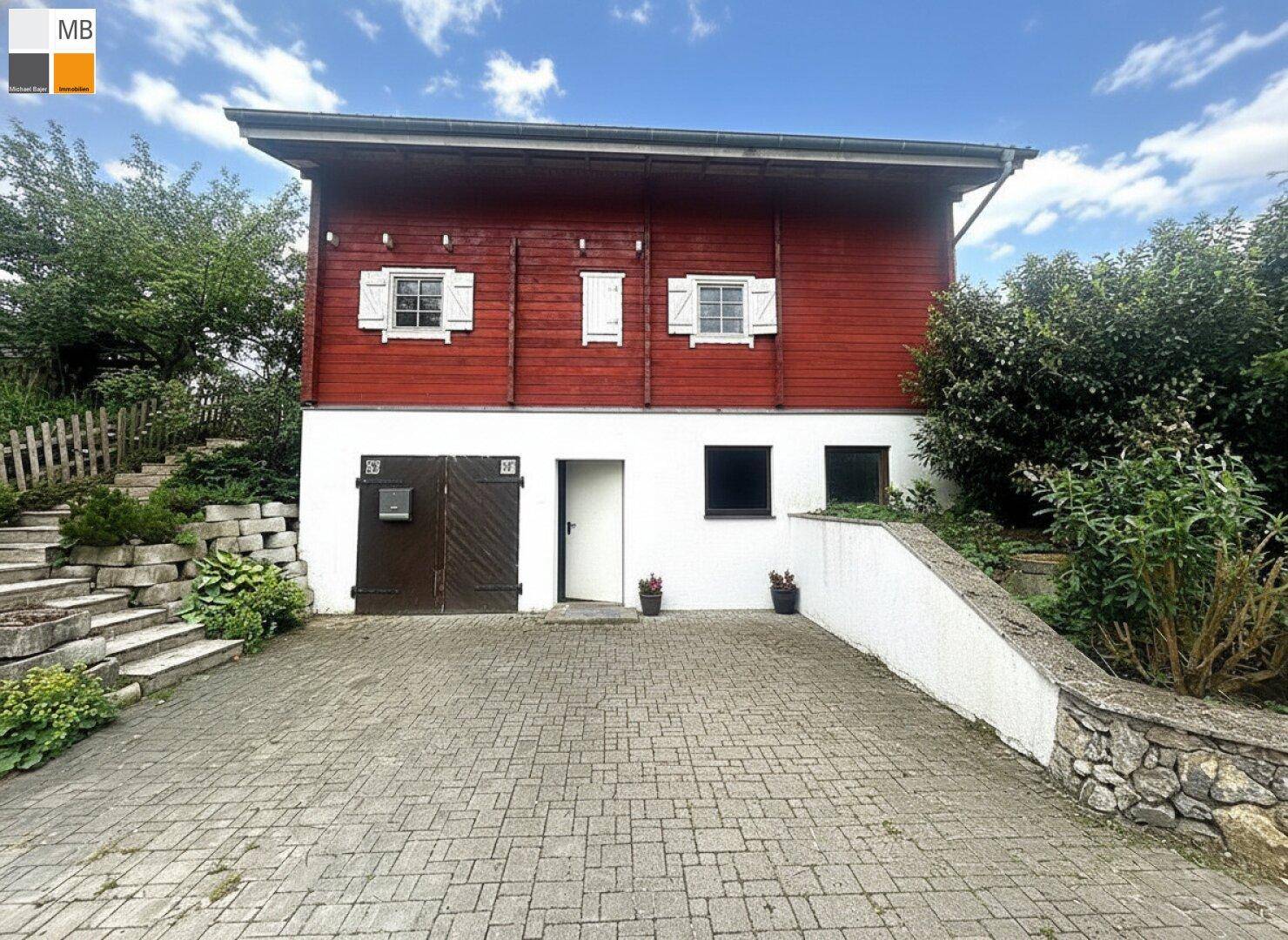 Vorderansicht des Hauses mit Garage und Eingangstür, gepflasterter Auffahrt und roter Holzfassade.