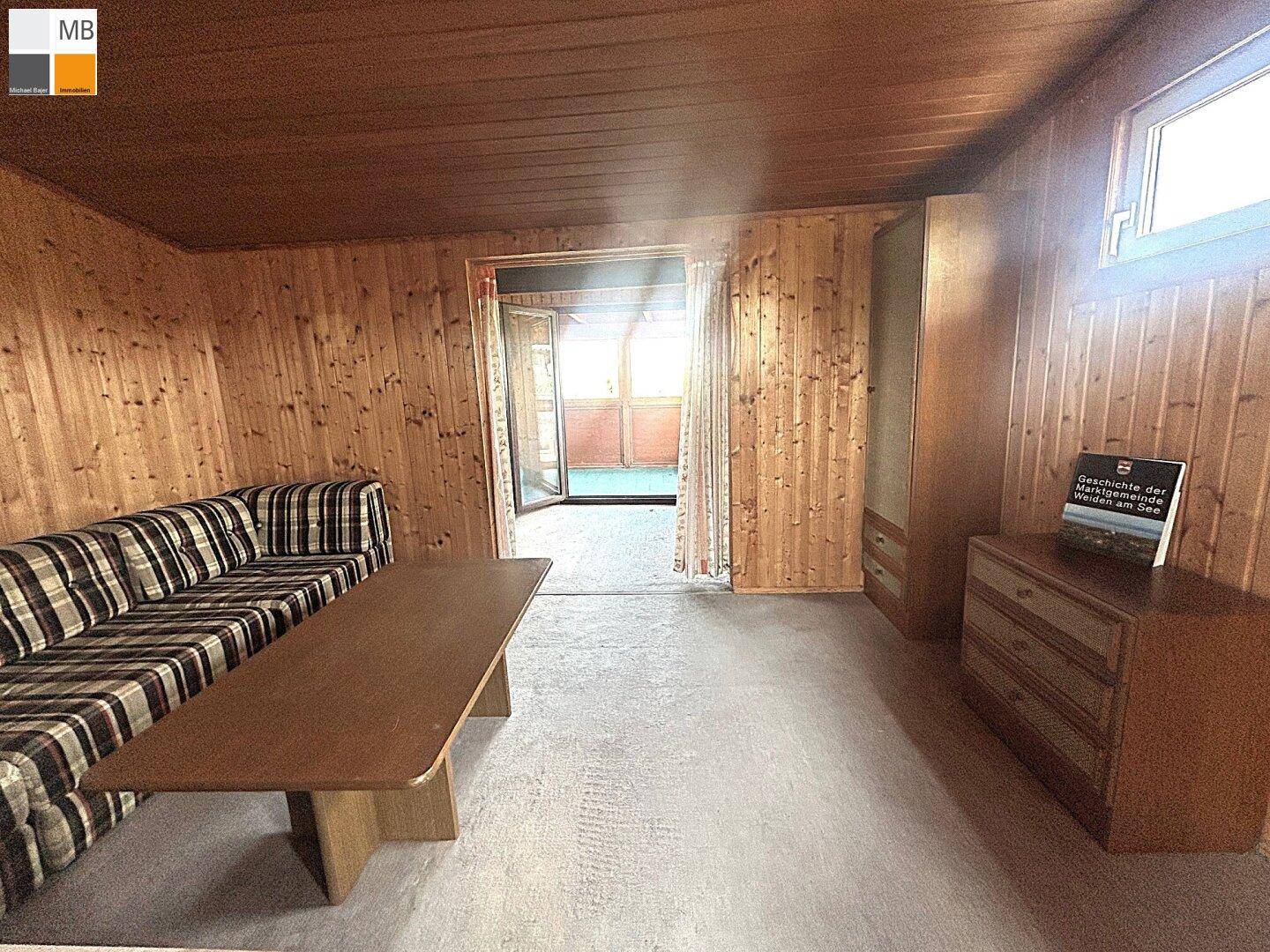 Helles Wohnzimmer mit Holzvertäfelung, Sofa und Zugang zu einem sonnigen Außenbereich.