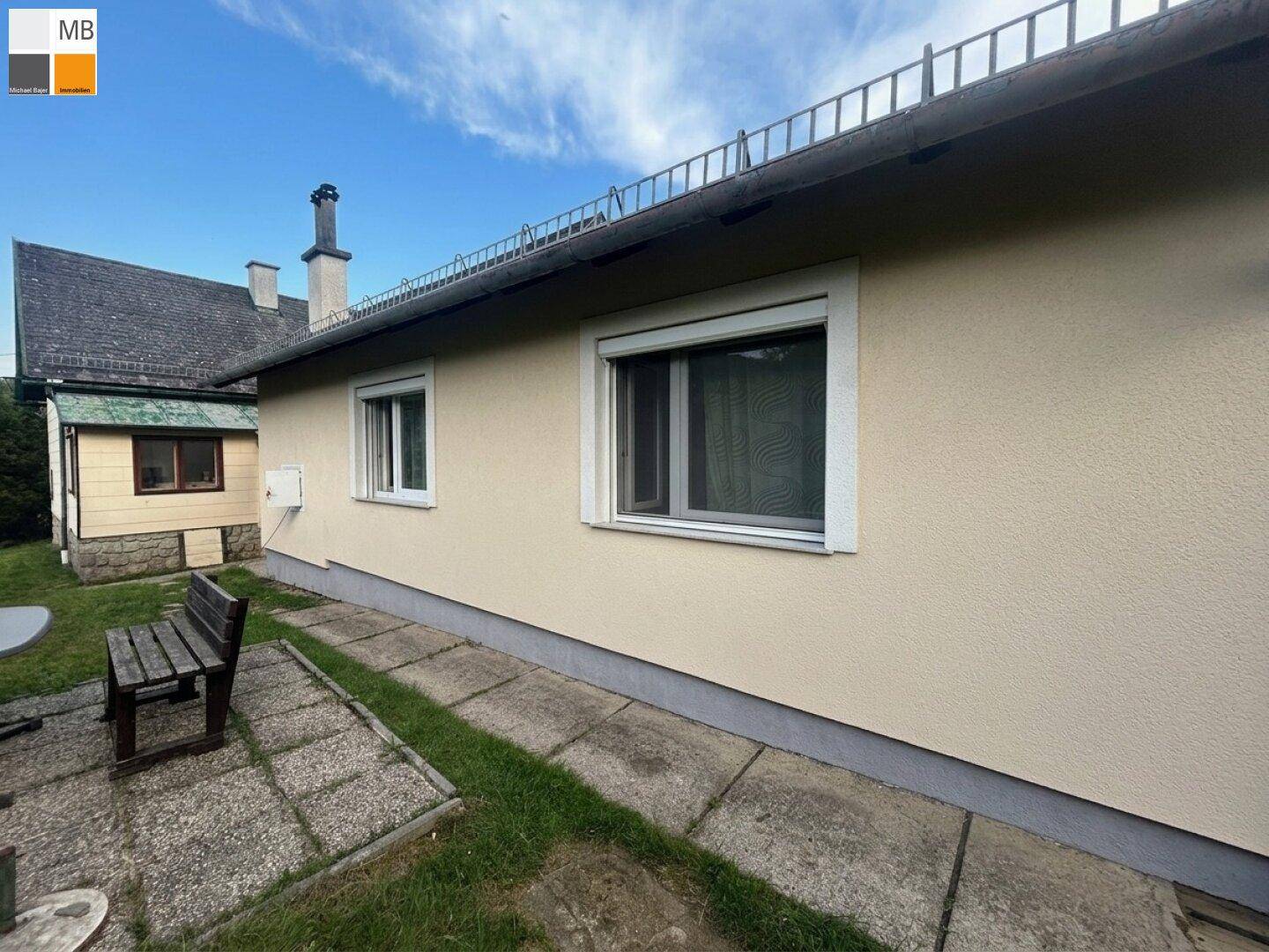 Seitenansicht des Hauses mit beigefarbener Fassade, Fenstern und einem kleinen Sitzbereich im Freien.