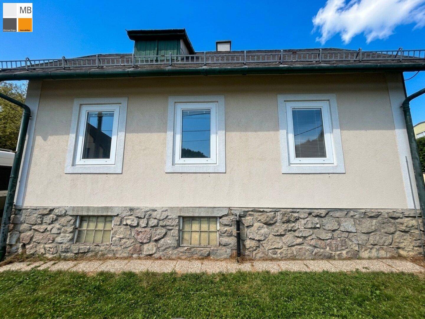 Rückansicht des Hauses mit Steinsockel, beigefarbener Fassade und drei Fenstern im Obergeschoss.