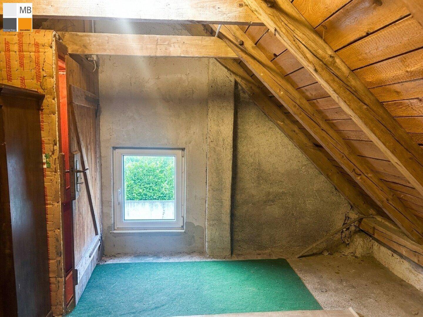 Ausgebauter Dachboden mit sichtbaren Holzbalken und einem Fenster mit Blick ins Grüne.