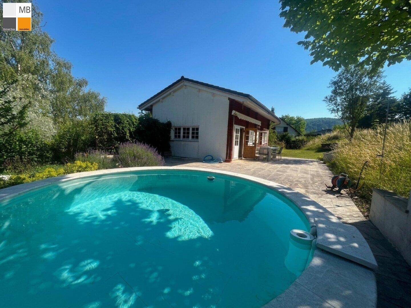 Runder Pool mit gepflasterter Terrasse und Blick auf das charmante Haus unter blauem Himmel.