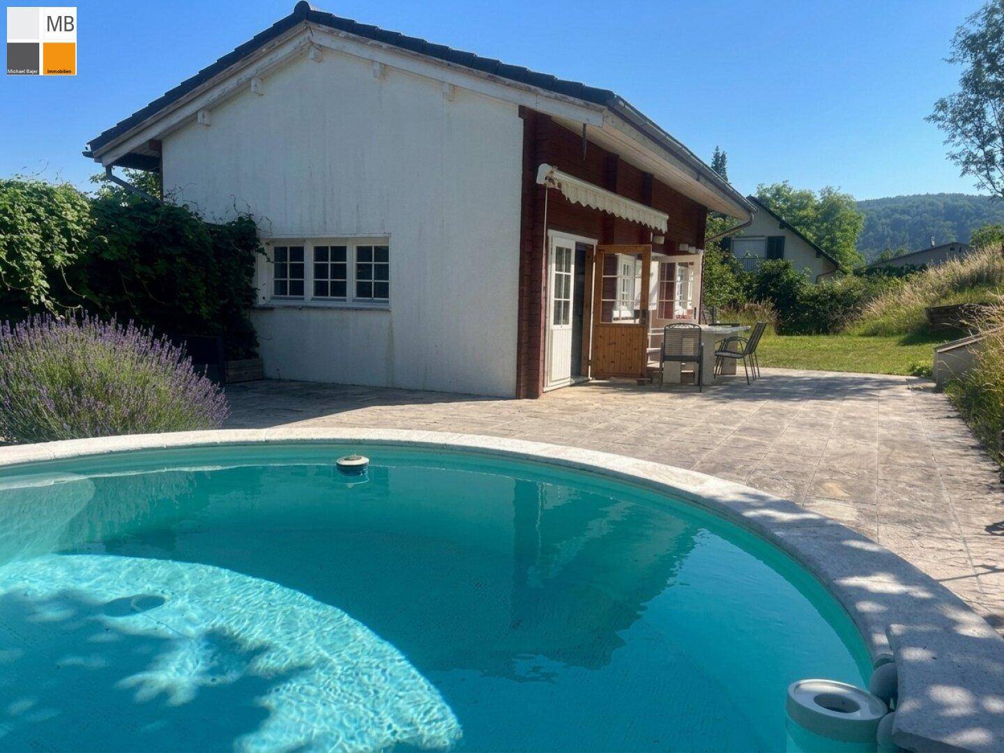 Runder Pool mit gepflasterter Terrasse und Blick auf das charmante Haus unter blauem Himmel.