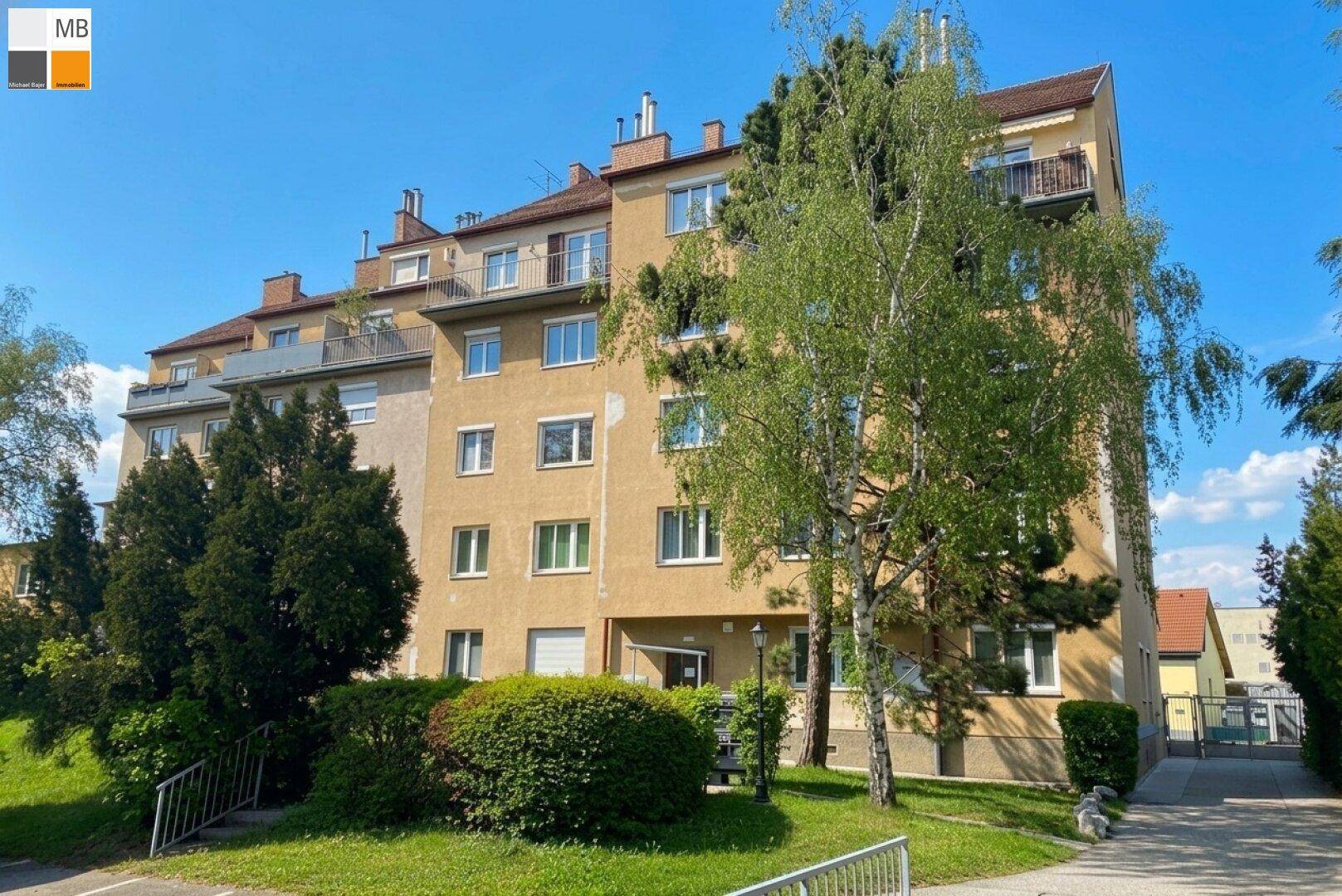 Mehrfamilienhaus mit Balkonen und grüner Umgebung unter blauem Himmel.