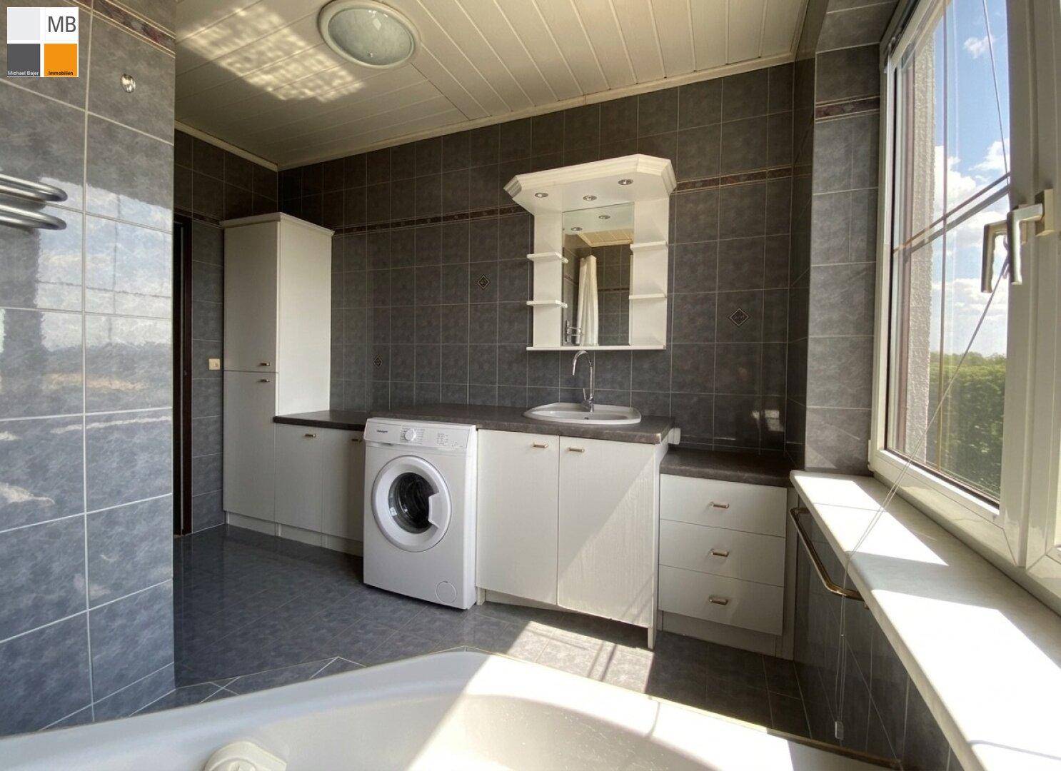 Badezimmer mit dunklen Fliesen, Waschbecken, Waschmaschine und Fenster.