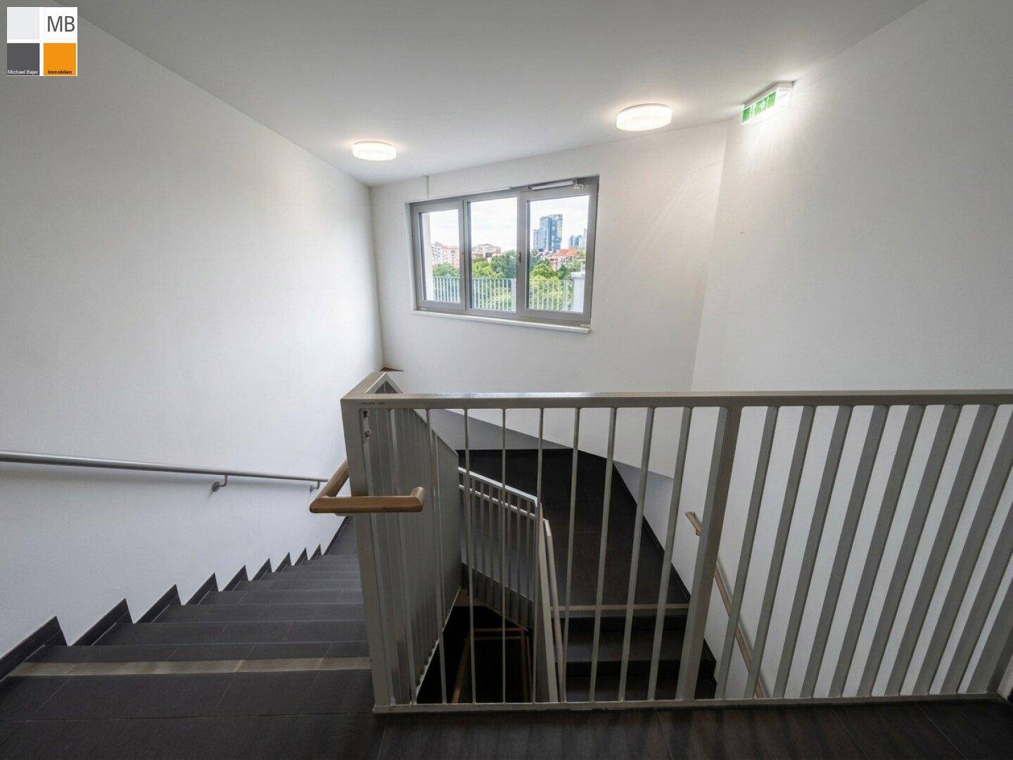 Treppenhaus mit modernem Geländer, großen Fenstern und Blick auf die Stadt.