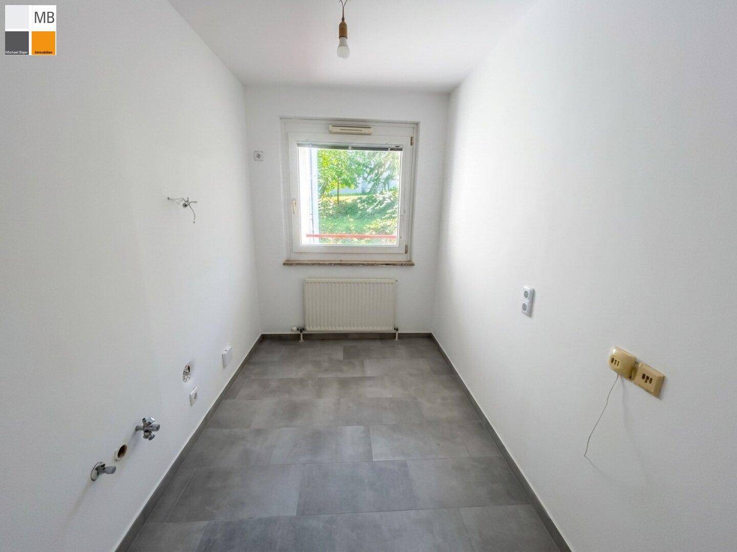 Leerer Raum mit grauen Fliesen, weißen Wänden und Fenster, vorbereitet für eine Kücheninstallation.