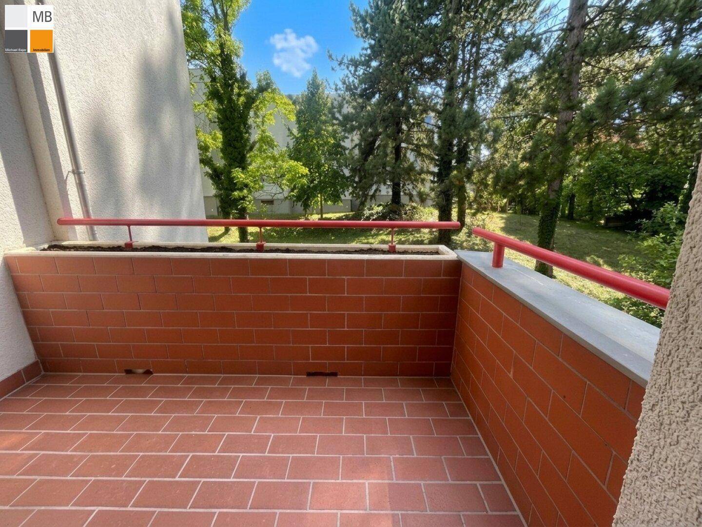 Geräumiger Balkon mit Terrakottafliesen und rotem Geländer, umgeben von viel Grün.