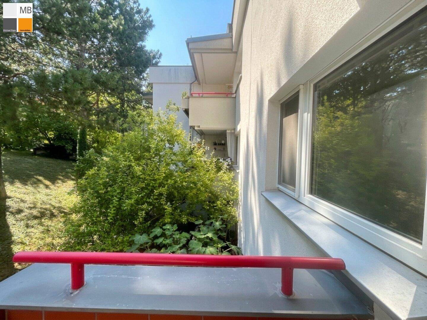 Seitlicher Blick vom Balkon auf die Gebäudefassade, Fenster und weitere Balkone mit grüner Umgebung.