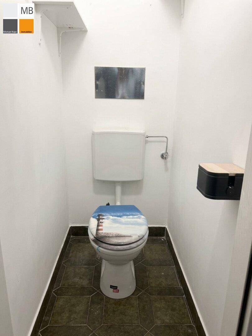 Separates WC mit weißen Wänden, dunklen Fliesen und einer Toilette mit dekorativem Sitz.