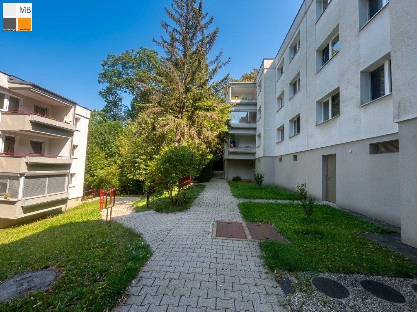 Außenansicht eines Wohnkomplexes mit gepflastertem Weg, grünen Rasenflächen und hohen Bäumen.