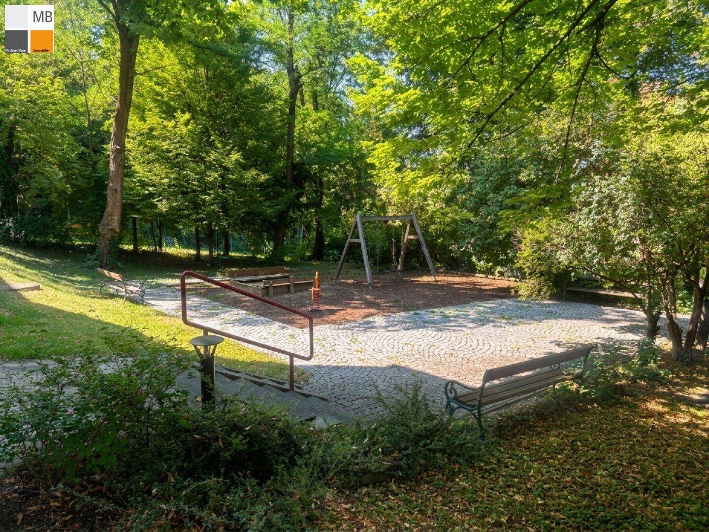 Öffentlicher Spielplatz mit Schaukeln und Bänken, umgeben von Bäumen und Grünflächen.