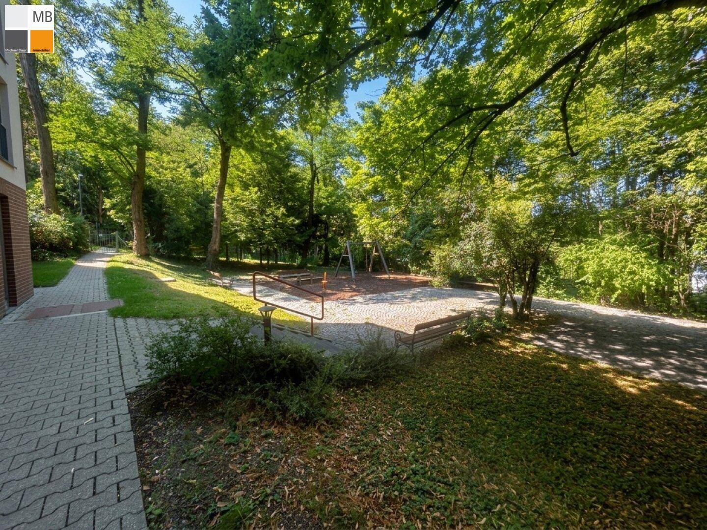 Blick auf den Spielplatz mit gepflastertem Zugangsweg, umgeben von üppigem Grün.