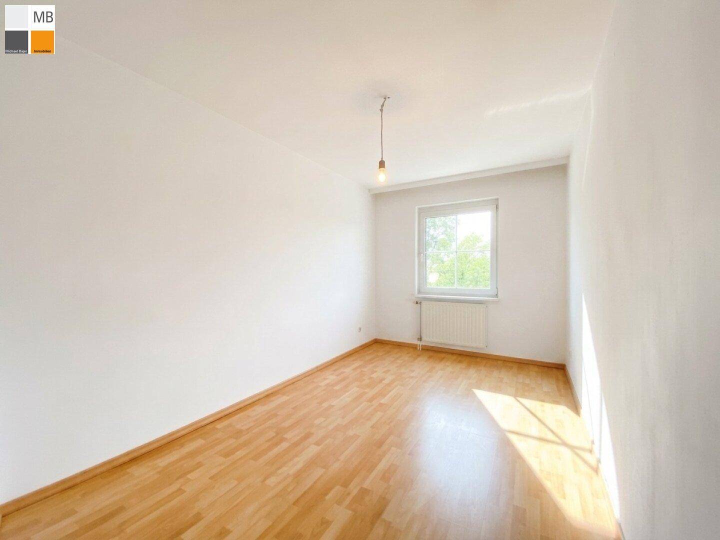 Leeres Zimmer mit hellem Laminatboden und einem Fenster, das den Raum mit Sonnenlicht füllt.