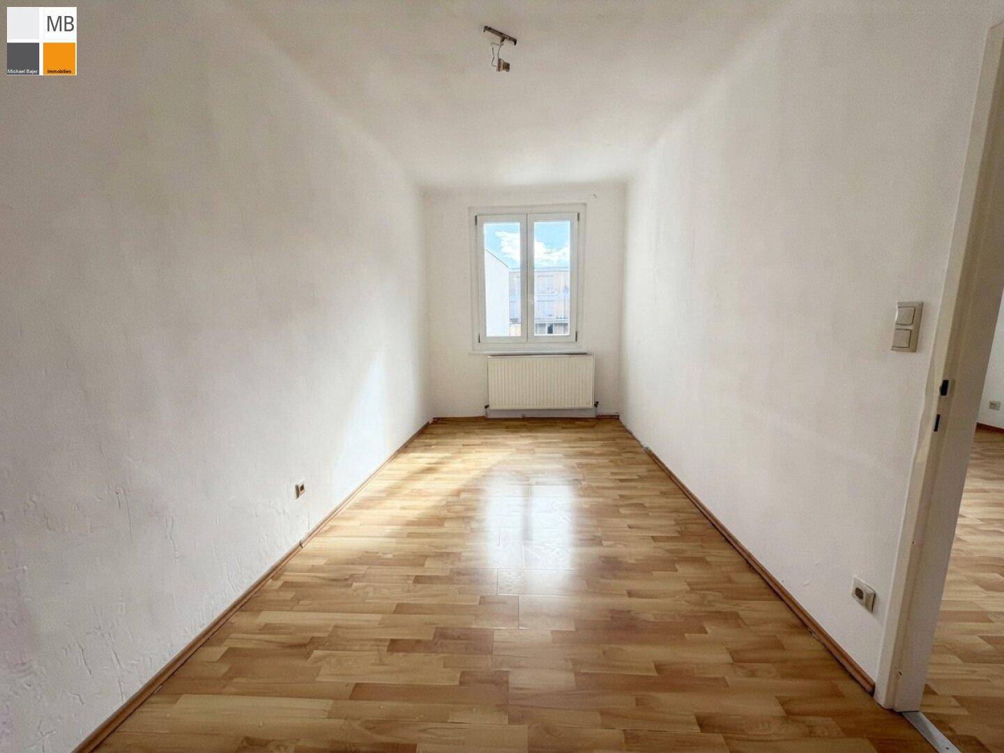 Leerer Raum mit Laminatboden, weißen Wänden und einem Fenster, das Tageslicht spendet.