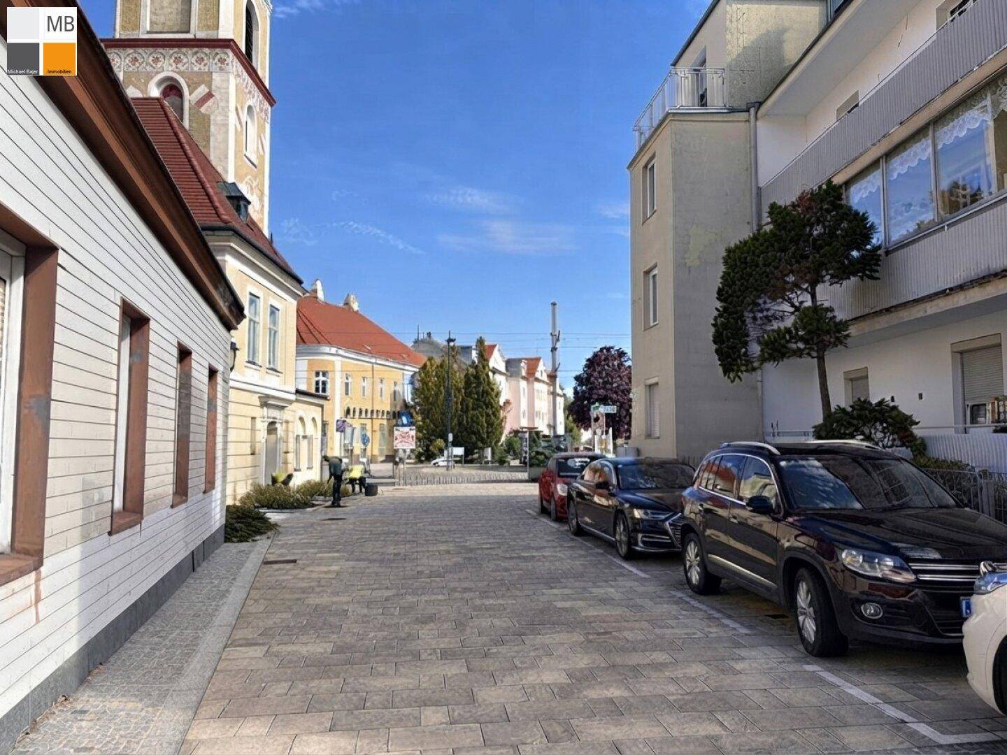 Historische Straße mit gepflastertem Boden, geparkten Autos und einem Kirchturm im Hintergrund.