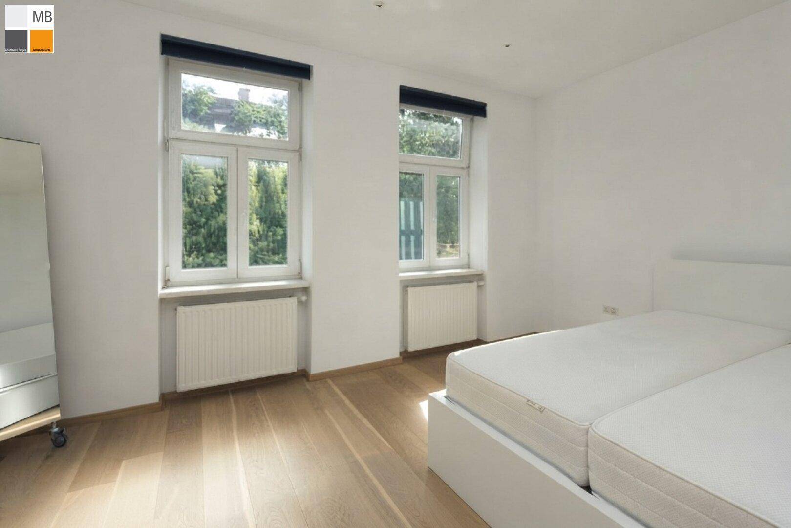 Helles Schlafzimmer mit Holzboden und zwei großen Fenstern, die viel Tageslicht hereinlassen.
