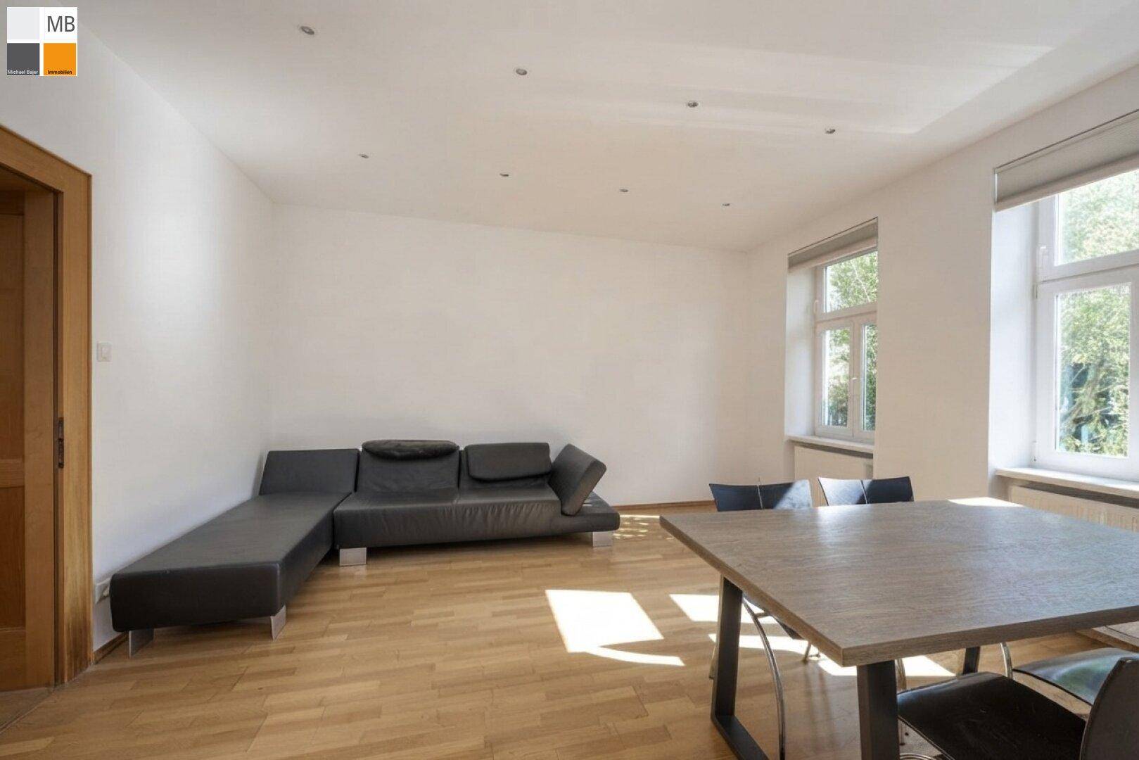 Geräumiges Wohnzimmer mit Holzboden, bequemer Couch und viel Tageslicht durch große Fenster.