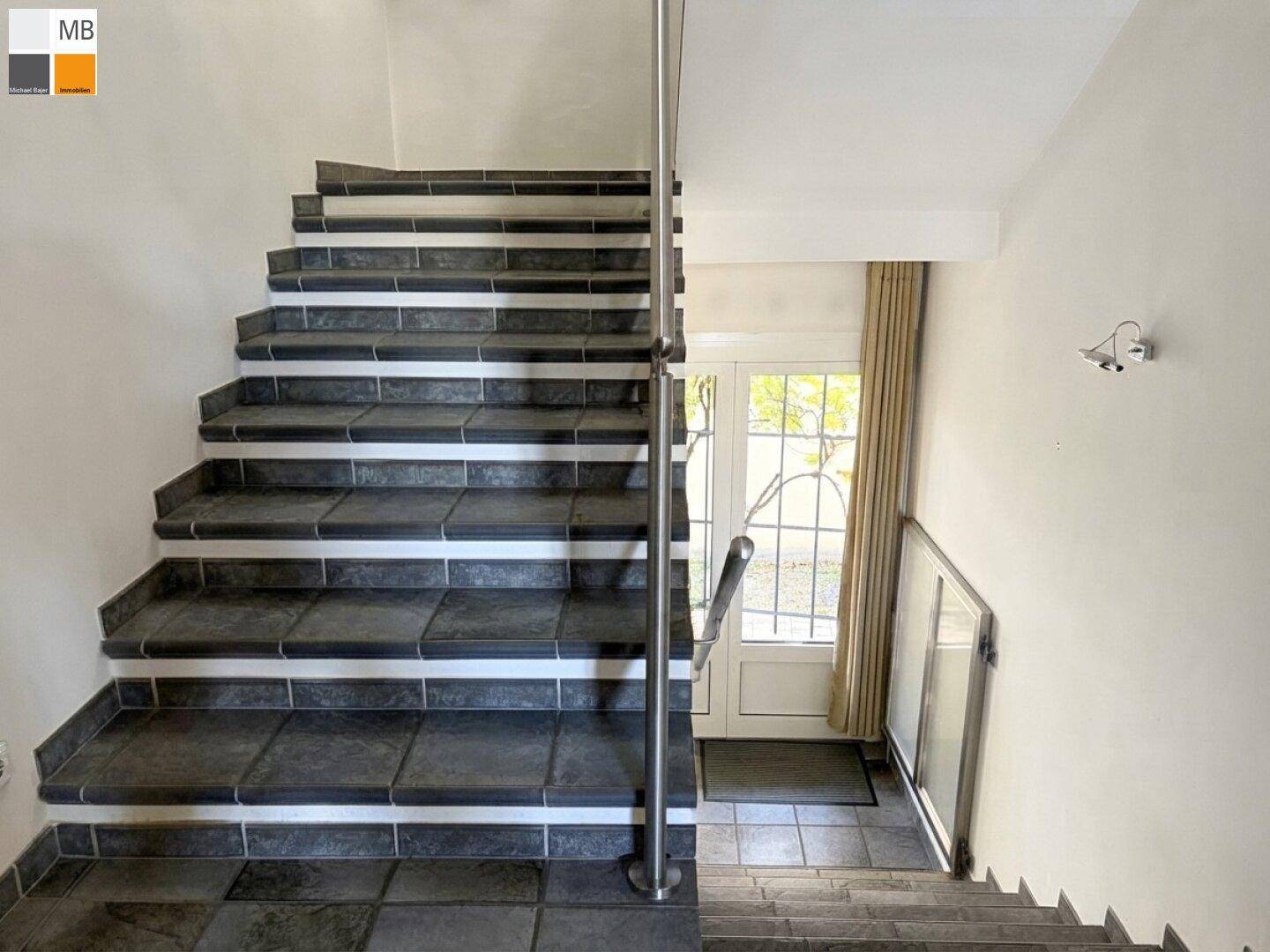 Treppenhaus mit dunklen Fliesenstufen und modernem Metallgeländer, führt zu den oberen Etagen.