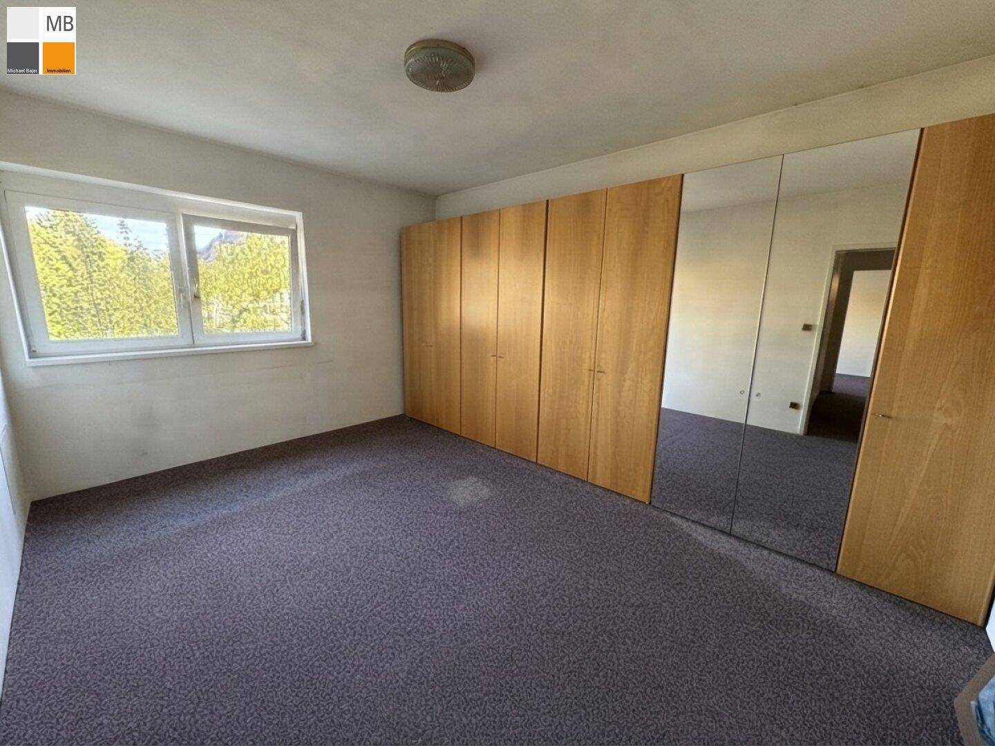 Schlafzimmer mit dunklem Teppichboden, eingebauten Holzschränken und einem Fenster mit Blick ins Grüne.