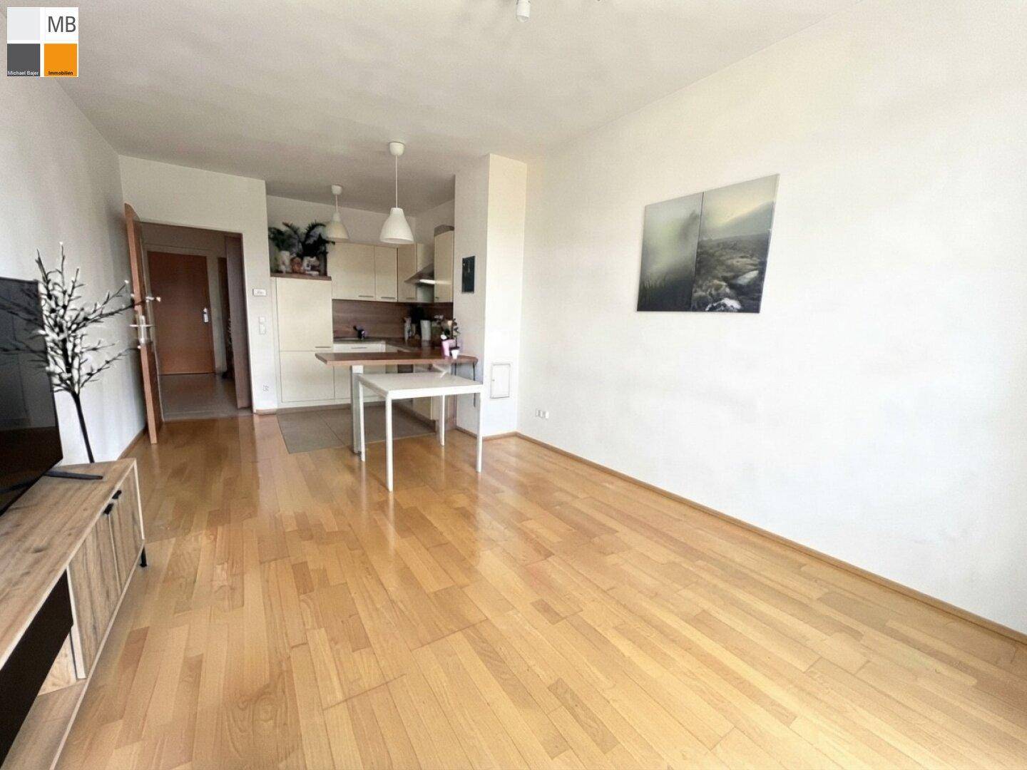 Wohnbereich mit hellem Parkettboden und integrierter Küchenzeile im Hintergrund, lichtdurchflutet.