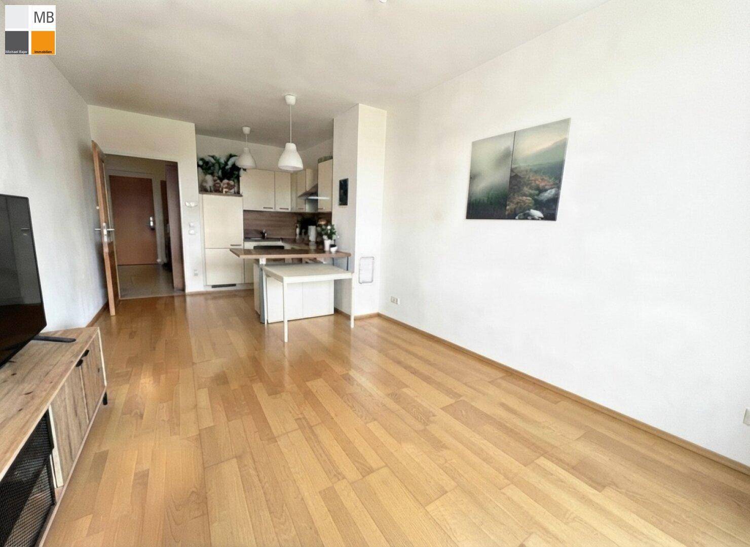 Heller Wohnbereich mit Holzboden und kleiner, funktionaler Küchenzeile im Hintergrund.
