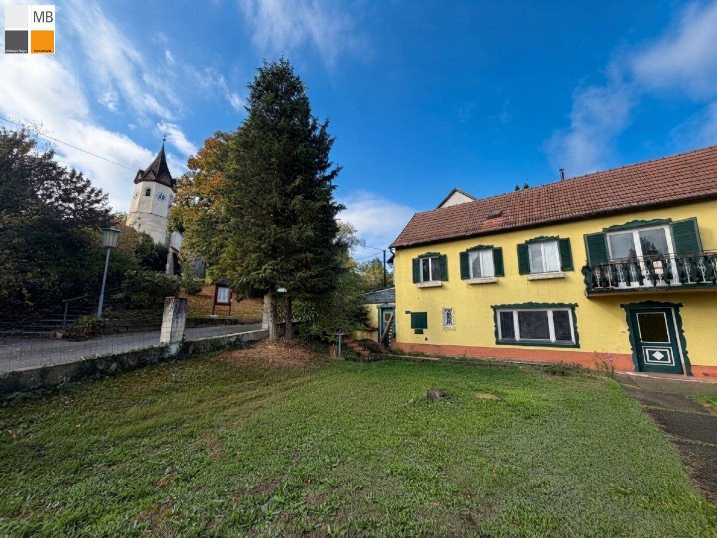 Gelbes Haus mit Garten und Blick auf einen historischen Kirchturm unter blauem Himmel.