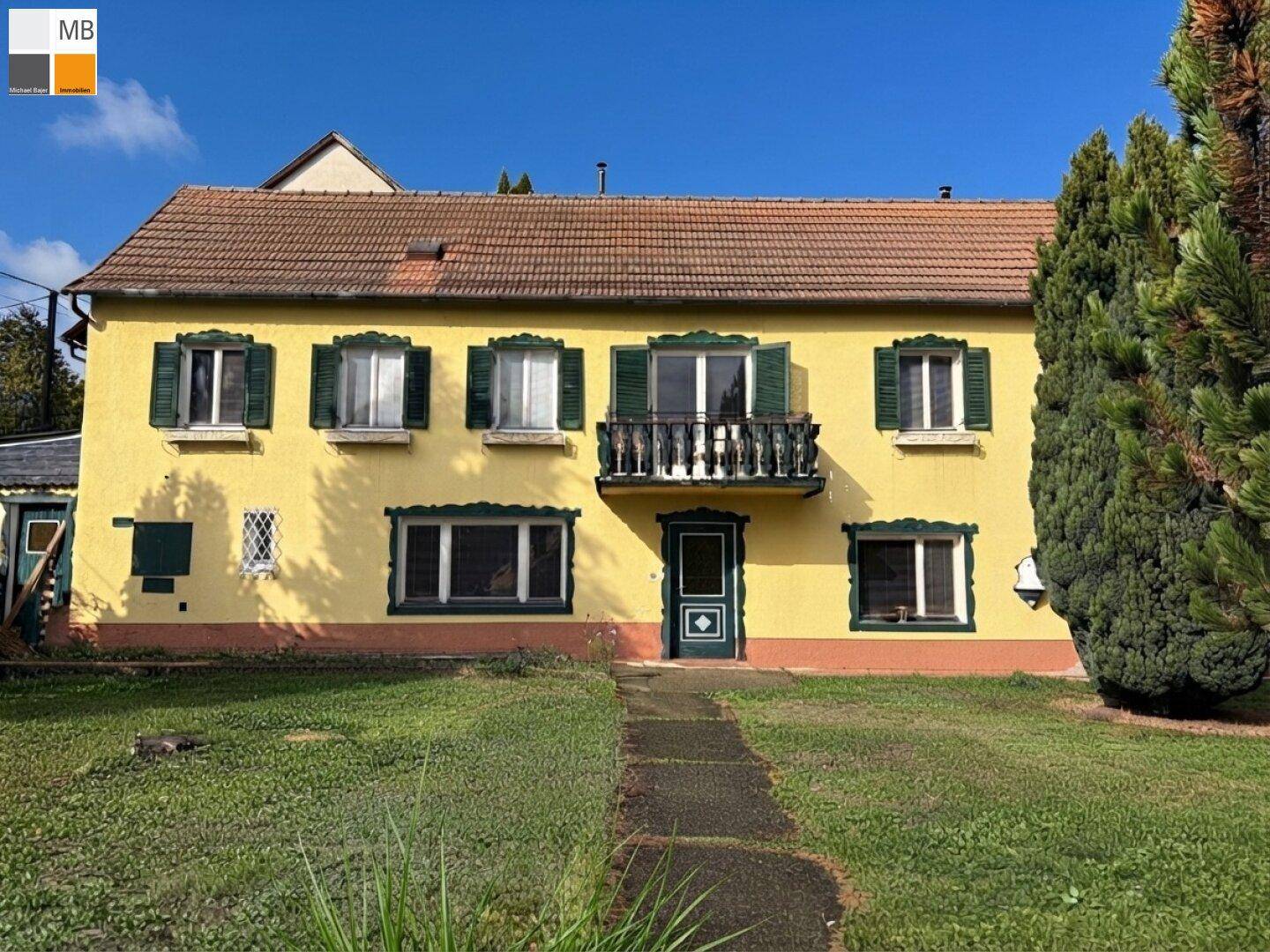 Frontansicht des gelben Hauses mit grünen Fensterläden, kleinem Balkon und gepflegtem Vorgarten.