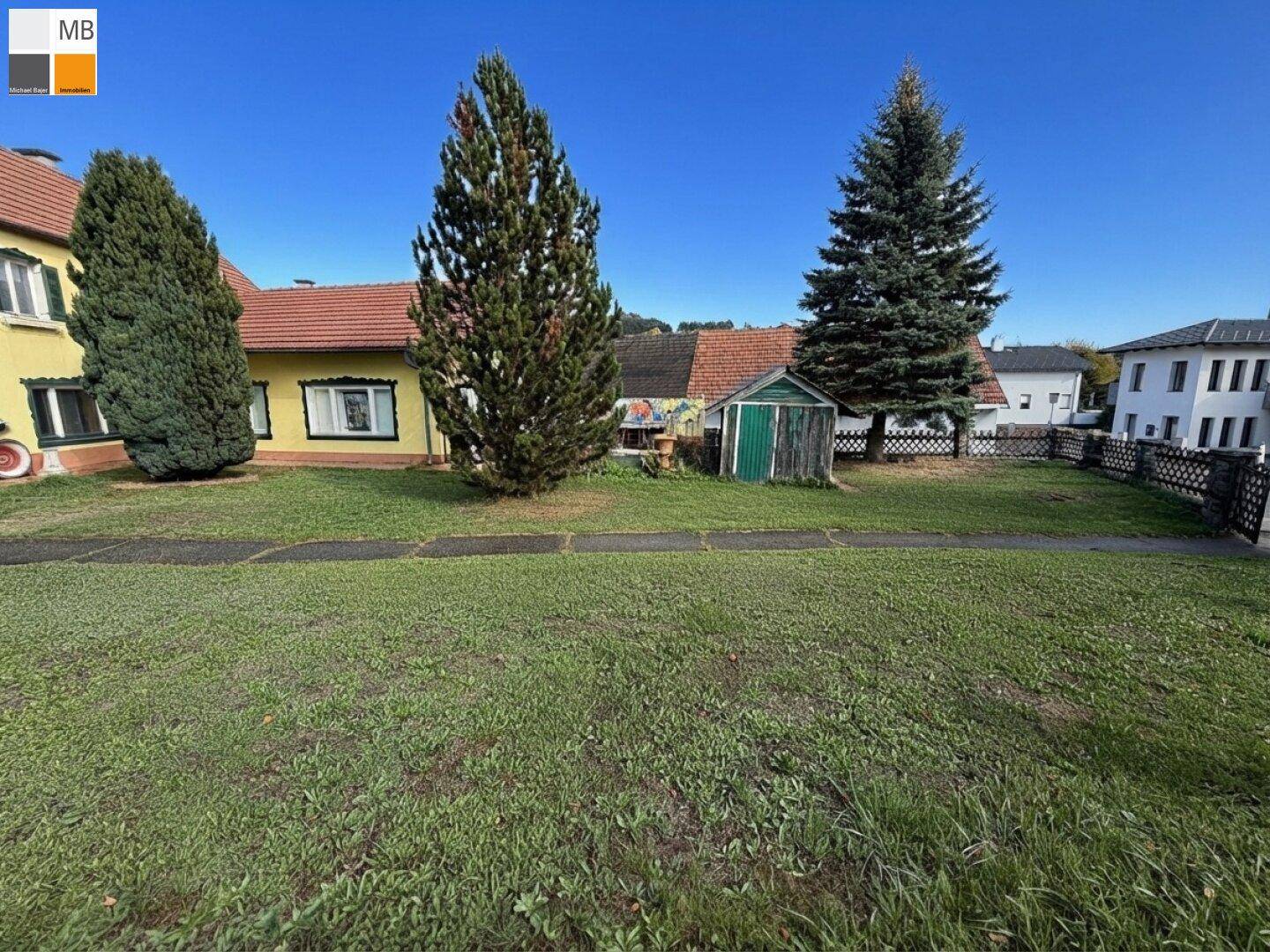 Ein gepflegter Gartenbereich mit Rasen, hohen Bäumen und einem kleinen Holzschuppen vor einem gelben Haus.