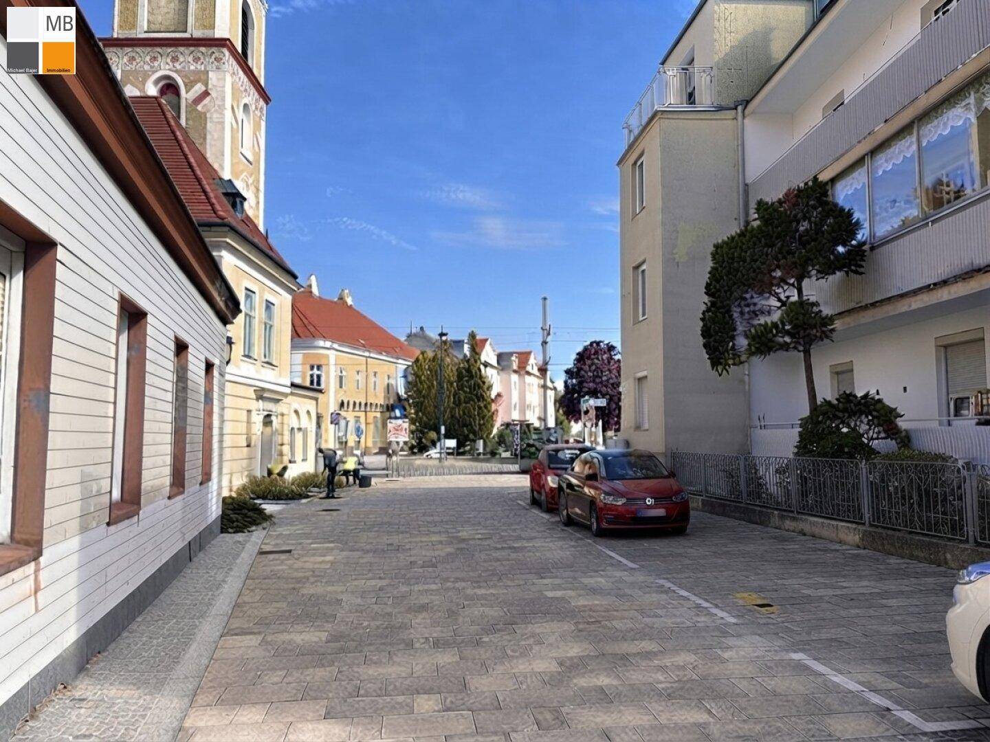 Historische Straße mit geparkten Autos, traditionellen Gebäuden und einem Kirchturm im Hintergrund.
