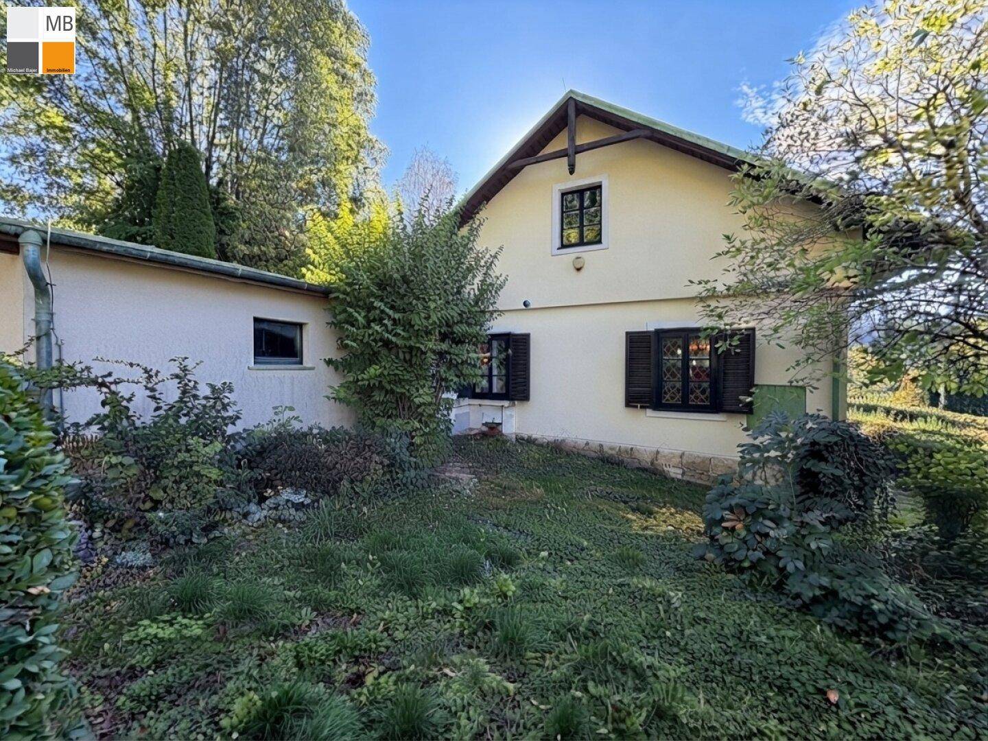 Rückseite des Hauses mit üppigem Grün und einem kleinen Gartenbereich unter strahlendem Sonnenschein.