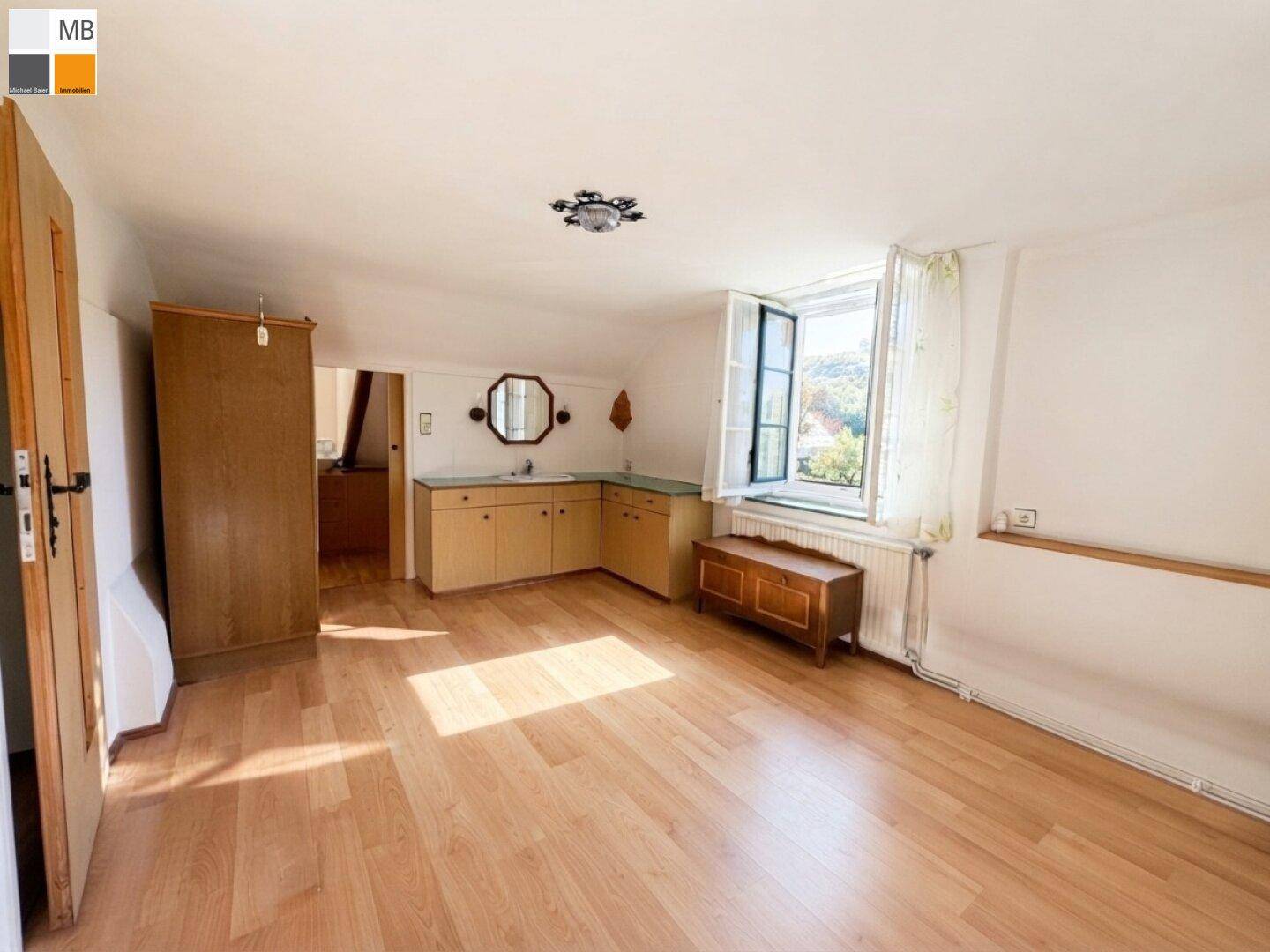Geräumiges Zimmer mit Laminatboden, kleiner Küchenzeile und Fenster mit Blick ins Grüne.