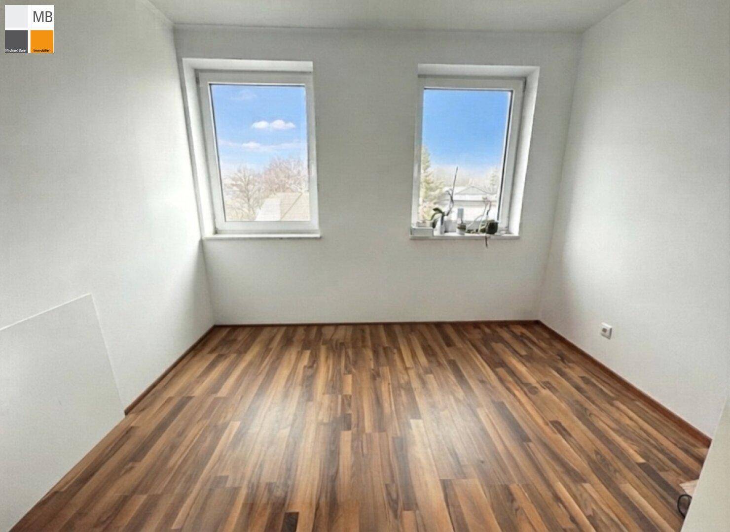 Kleines Zimmer mit Holzboden und zwei Dachfenstern, die den Raum erhellen.