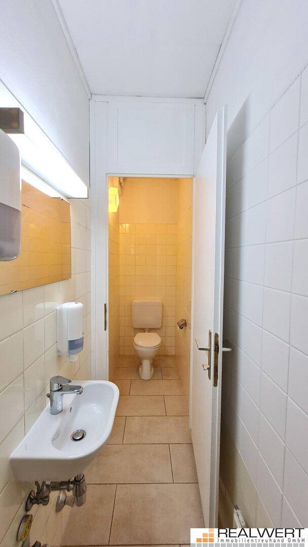 Kleiner Toilettenraum mit Waschbecken, Spiegel und hellen Fliesen an den Wänden.