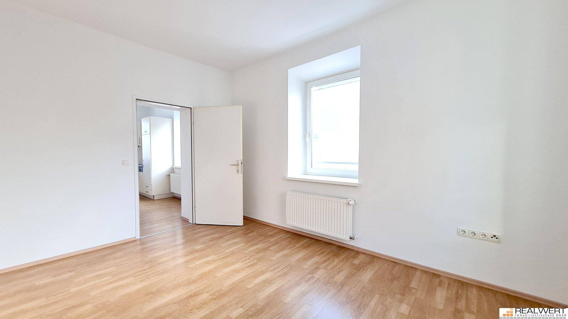 Heller, leerer Raum mit Laminatboden, großem Fenster und Heizkörper unter dem Fenster.