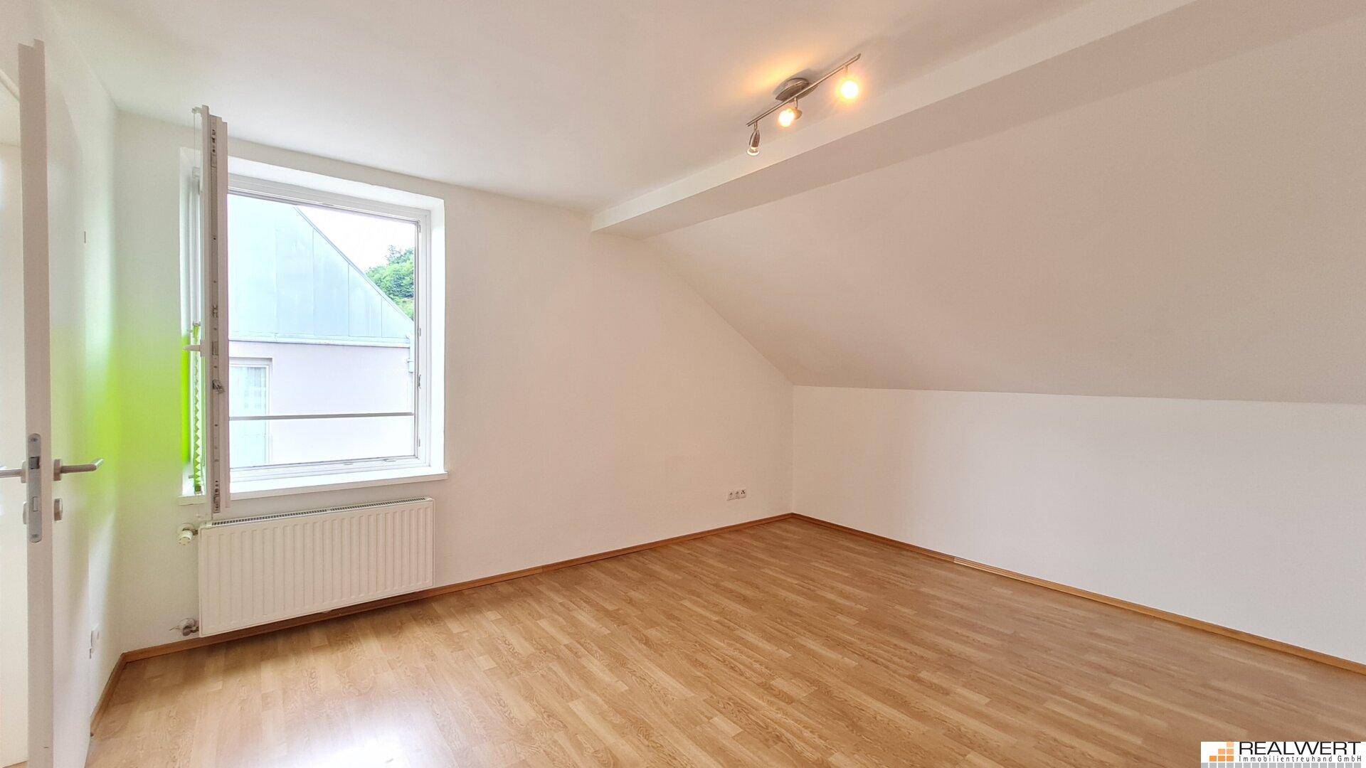 Leerer Raum im Dachgeschoss mit Laminatboden, Fenster und schrägen Wänden.