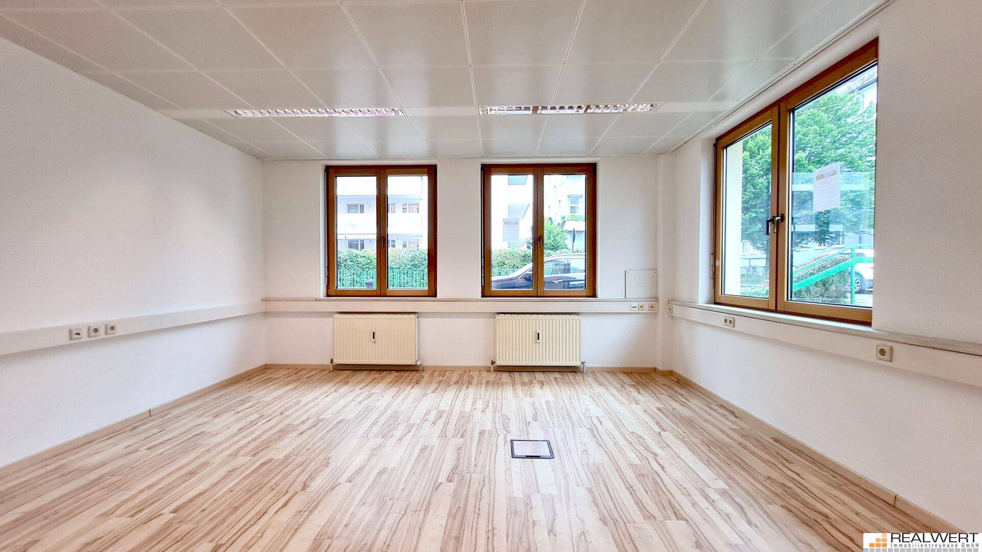 Heller, leerer Büroraum mit großen Fenstern, hellem Laminatboden und abgehängter Decke.