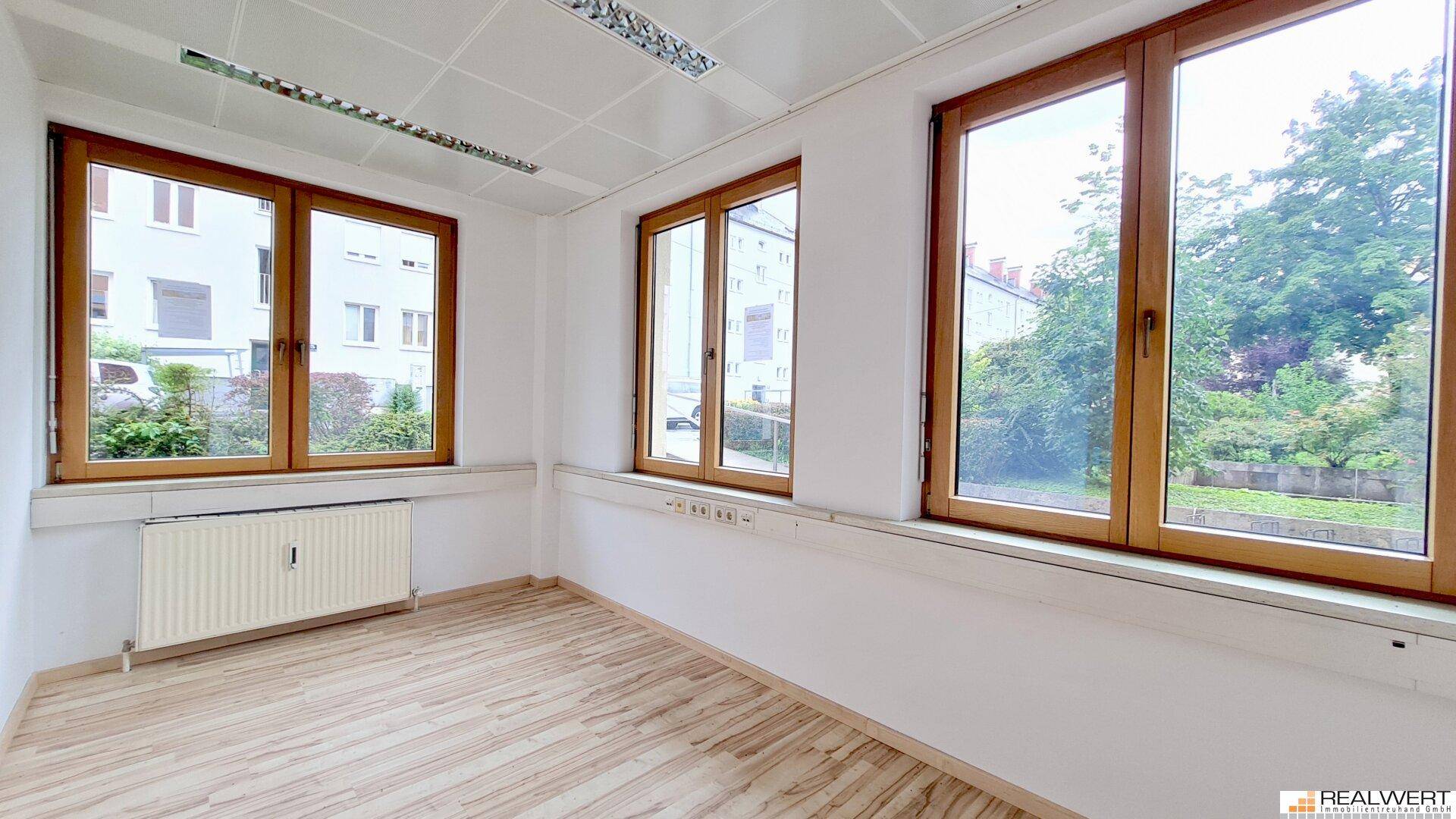Geräumiger Büroraum mit mehreren Fenstern, die viel Tageslicht hereinlassen und einen Blick ins Grüne bieten.