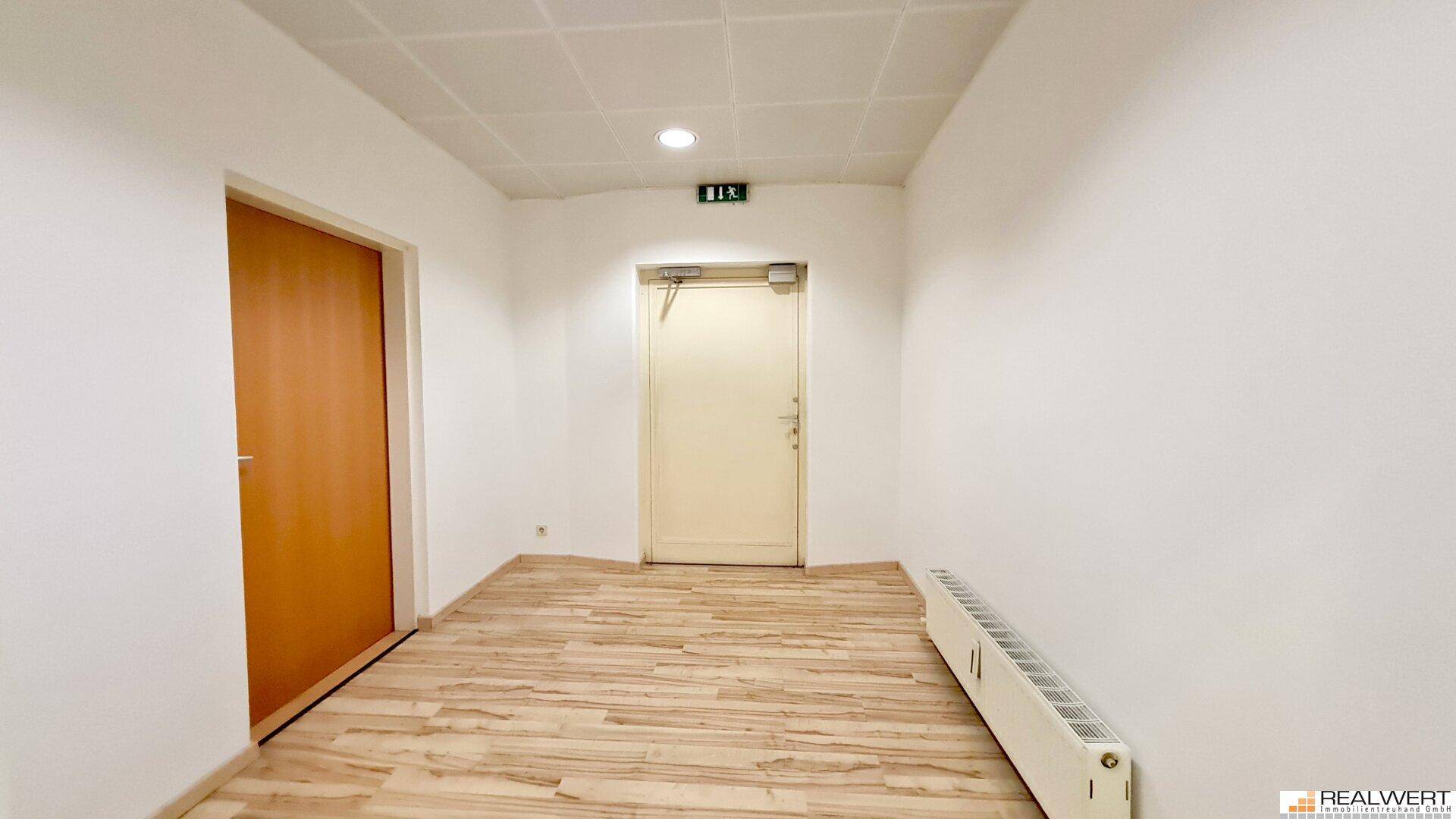 Heller Flur mit Laminatboden, weißen Wänden und zwei Türen, eine davon ein Notausgang.