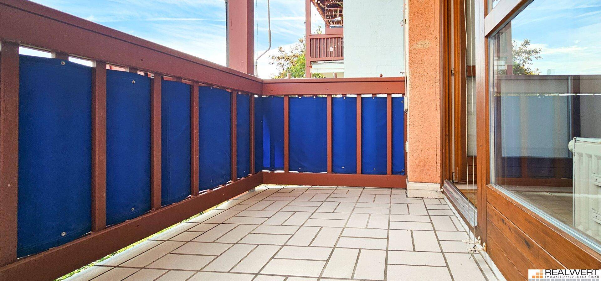 Geräumiger Balkon mit Fliesenboden, Holzgeländer und blauen Sichtschutzwänden.