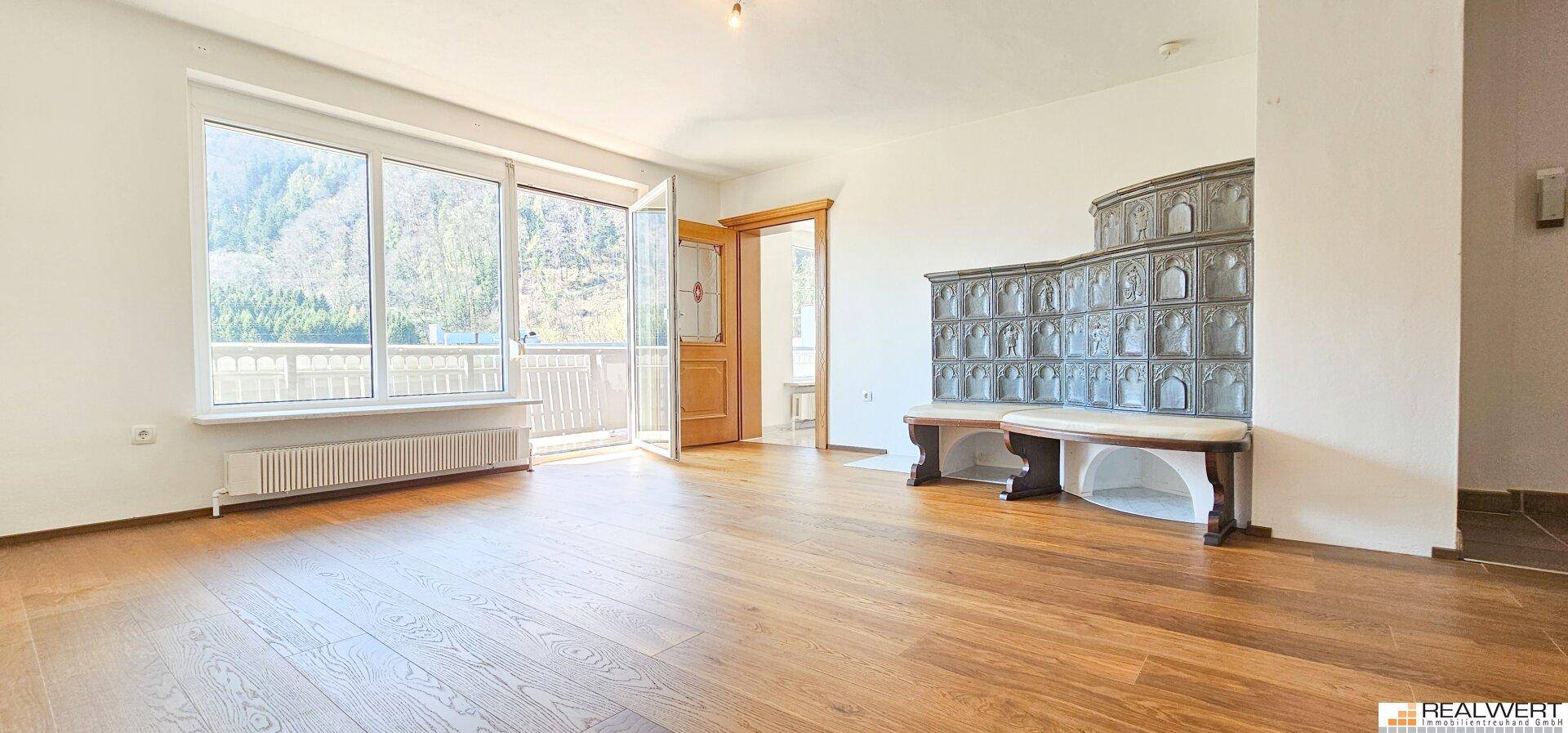 Geräumiges Wohnzimmer mit Holzboden, großem Kachelofen und Zugang zu einem Balkon.