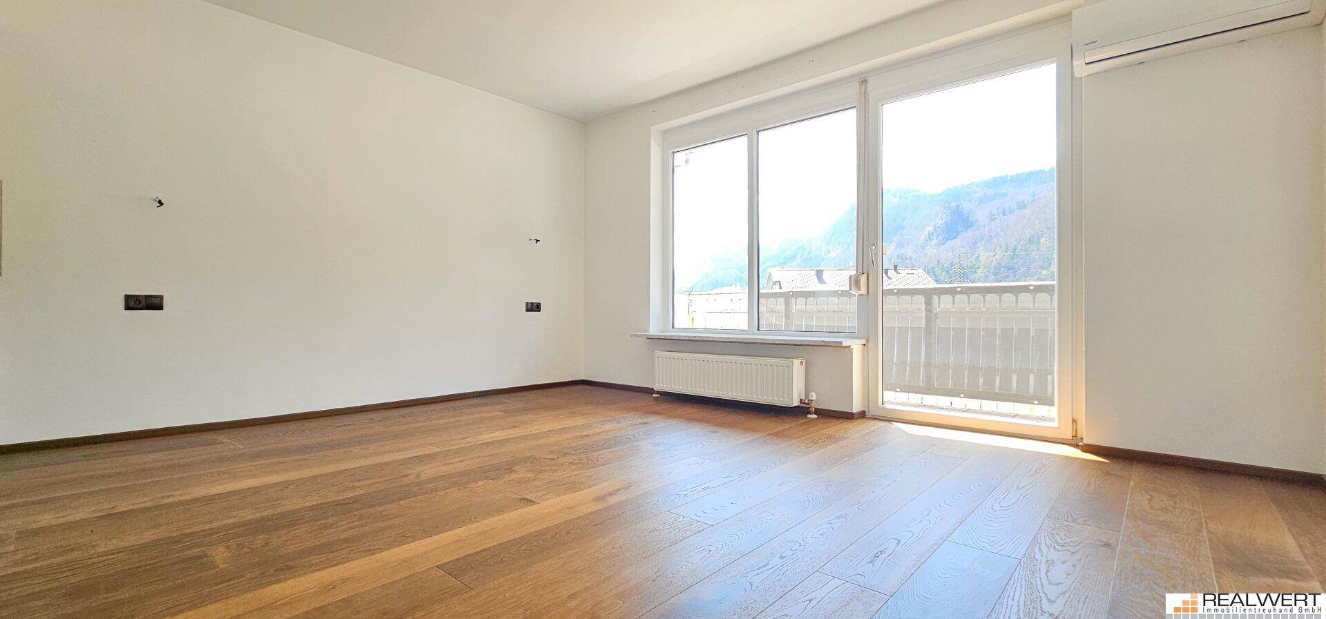 Heller Raum mit Holzboden, großen Fenstern und Zugang zu einem Balkon mit Bergblick.