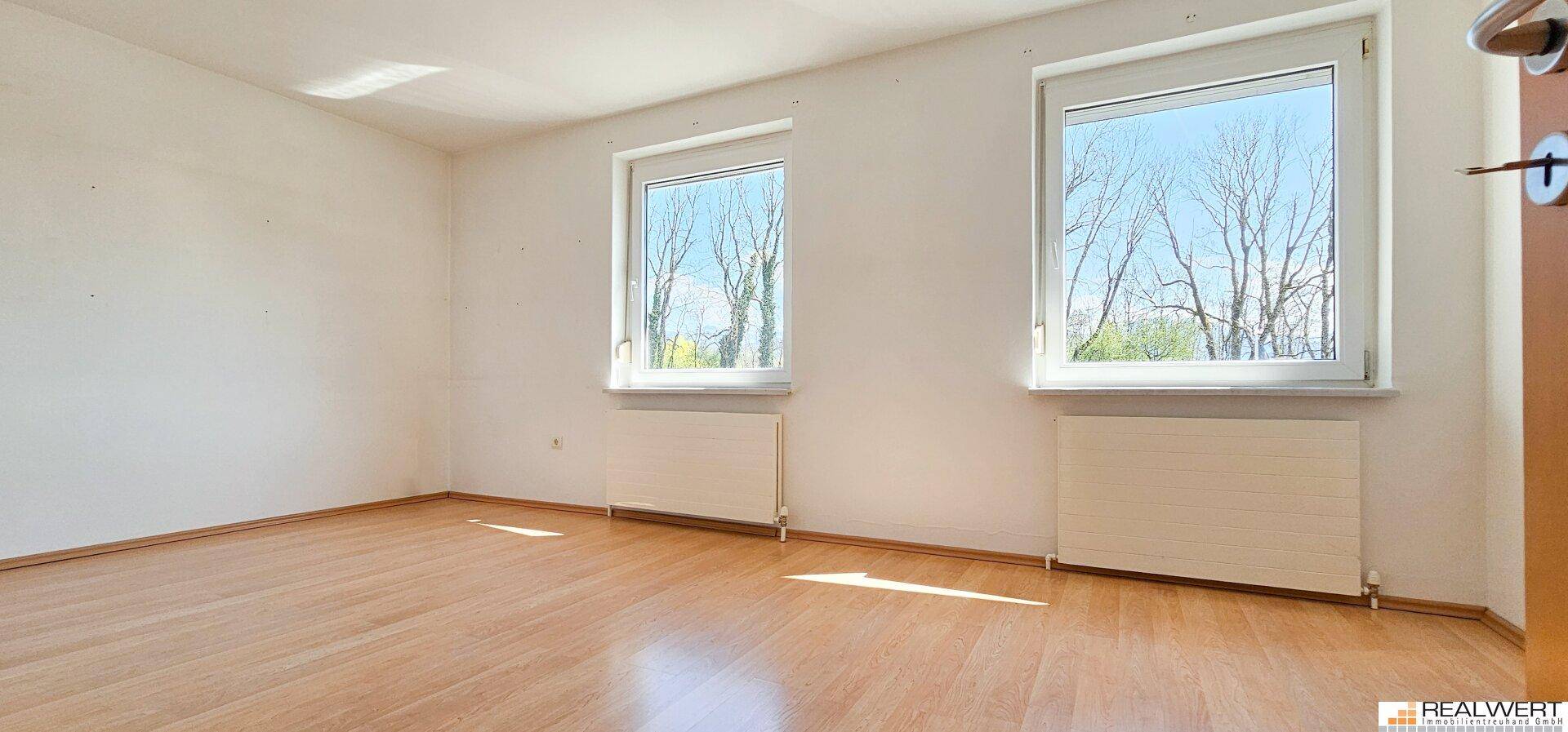 Helles Schlafzimmer mit Holzboden, weißen Wänden und zwei Fenstern mit Blick ins Grüne.