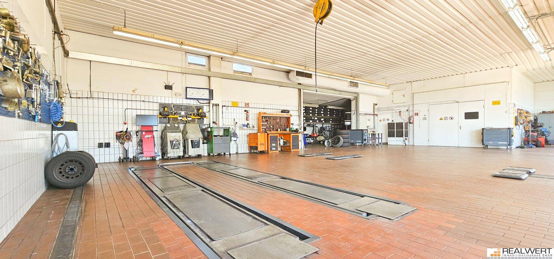 Weitläufige Werkstatt mit Fliesenboden, verschiedenen Arbeitsstationen und einem Reifenlager im Hintergrund.