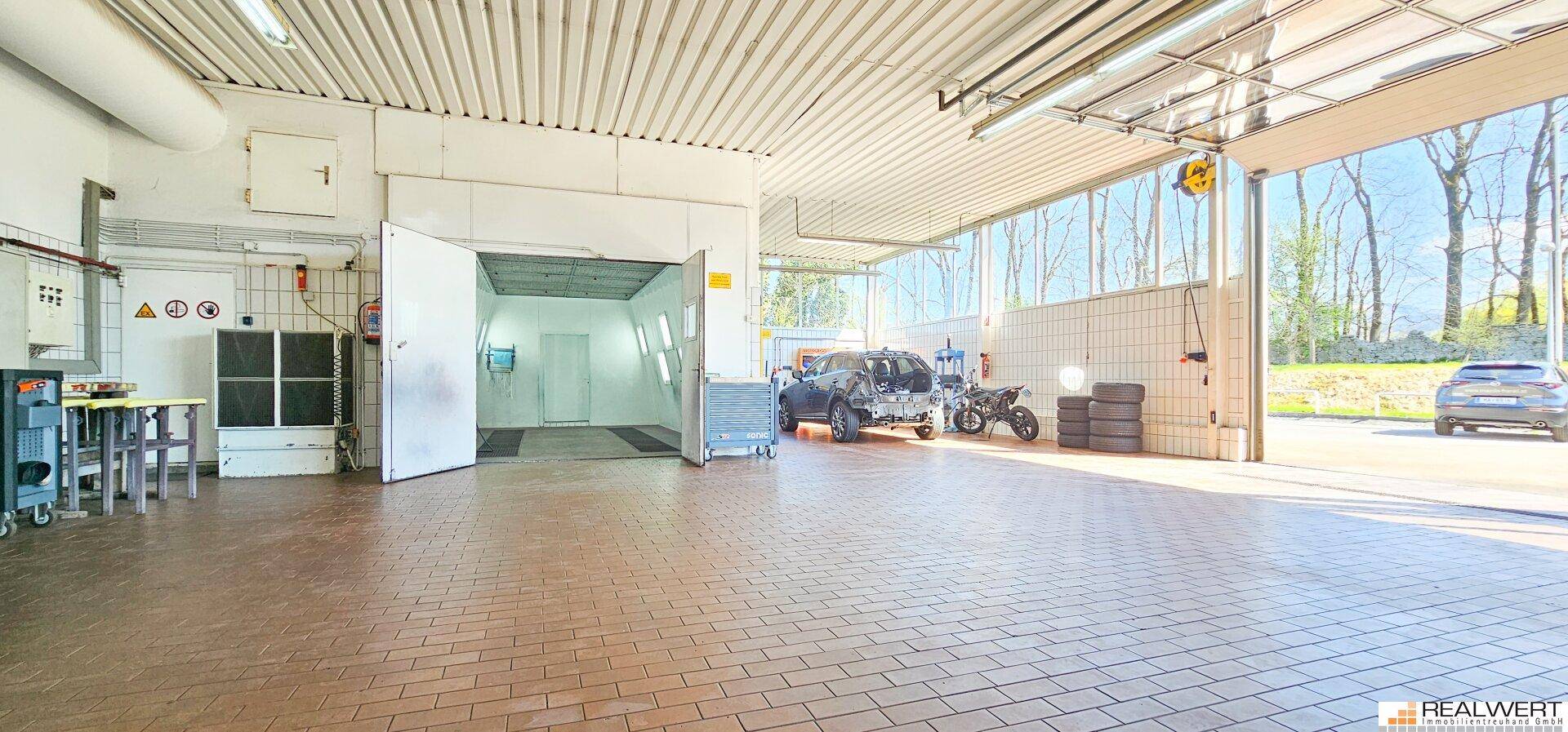 Autowerkstatt mit geöffneter Lackierkabine, Fliesenboden und Tageslicht durch große Fenster.
