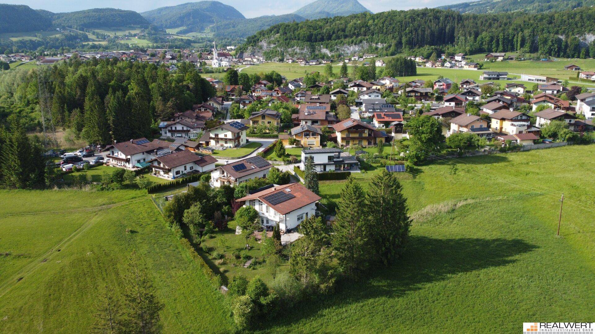 Weitläufige Luftaufnahme der Siedlung mit Häusern, grünen Feldern und einer malerischen Bergkulisse.
