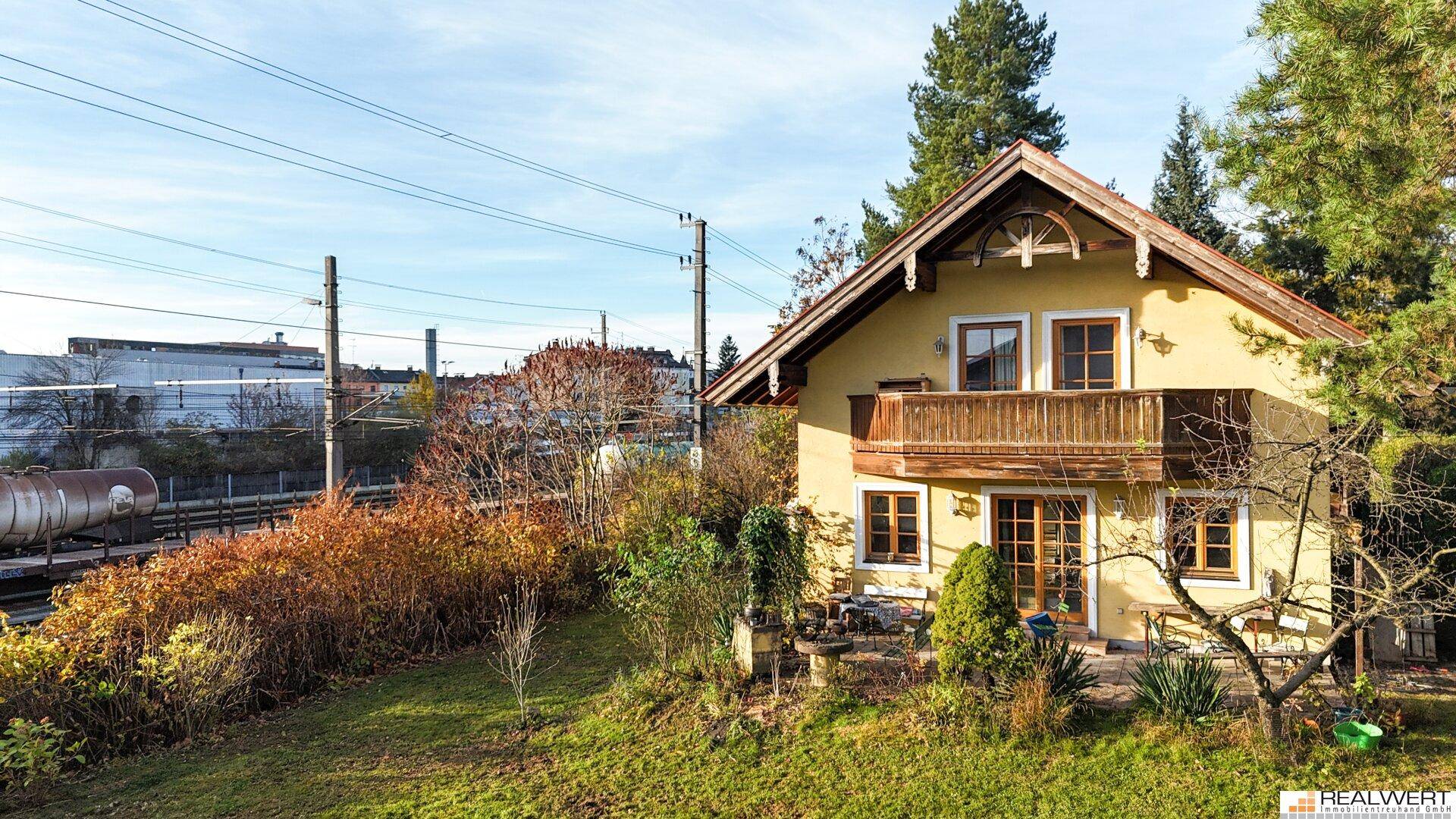 Traditionelles Haus mit gelber Fassade und Holzbalkon, angrenzend an Bahngleise und grüne Landschaft.