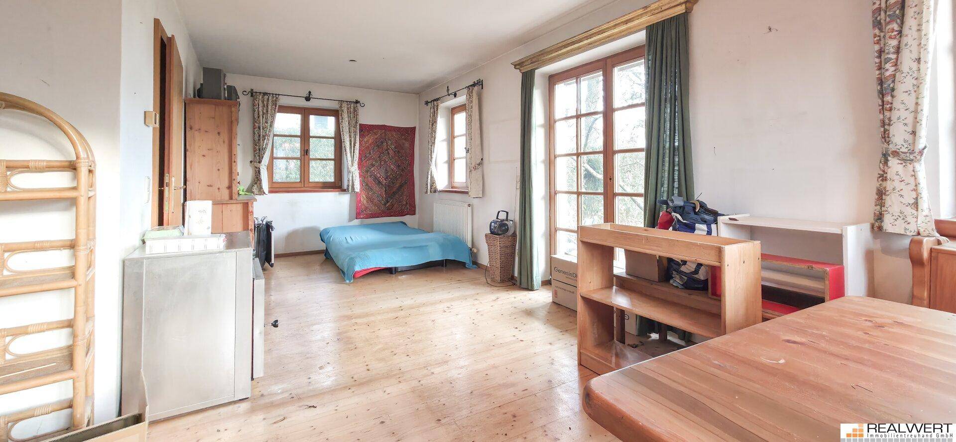 Geräumiges Zimmer mit Holzboden, großen Fenstern und einfacher Einrichtung, ideal als Schlaf- oder Wohnbereich.